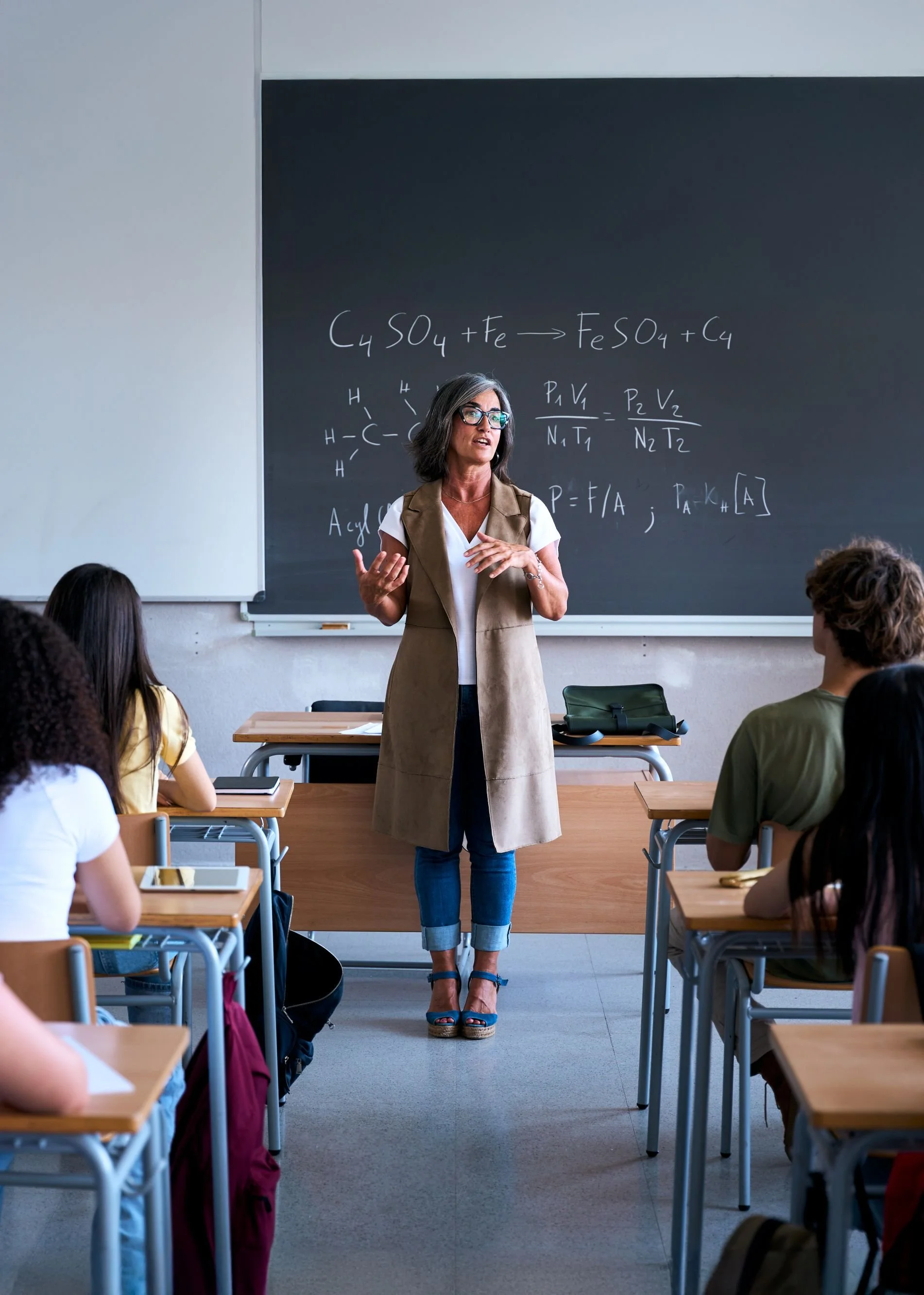 A teacher standing in front of a chalkboard in a classroom, giving a lecture to students. The chalkboard has chemical equations and formulas written on it.