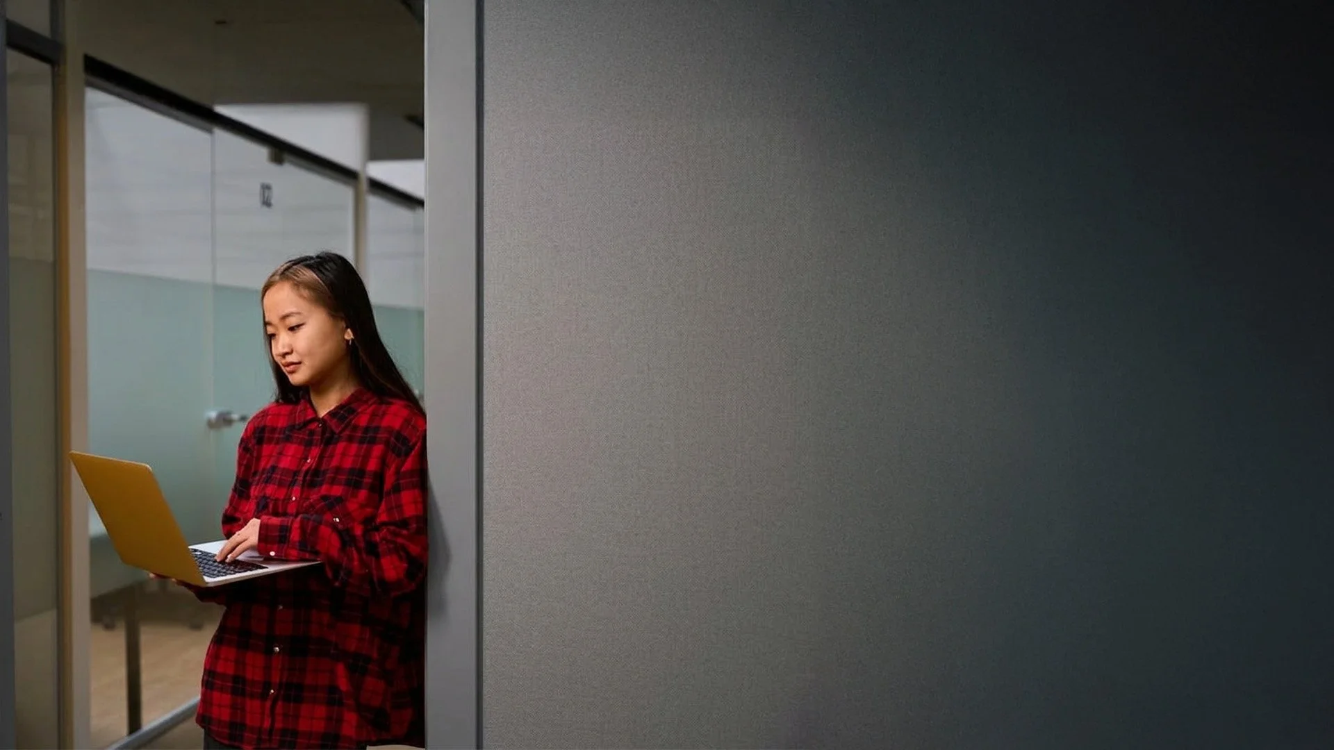 A young woman with long dark hair wearing a red and black plaid shirt, standing in a modern office corridor, looking down at her yellow laptop.