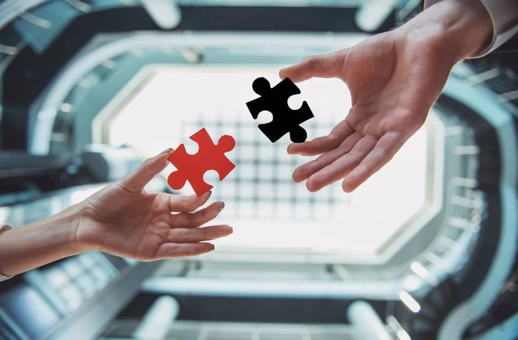Two hands exchanging puzzle pieces, one red and one black, with a modern architectural background and natural light.