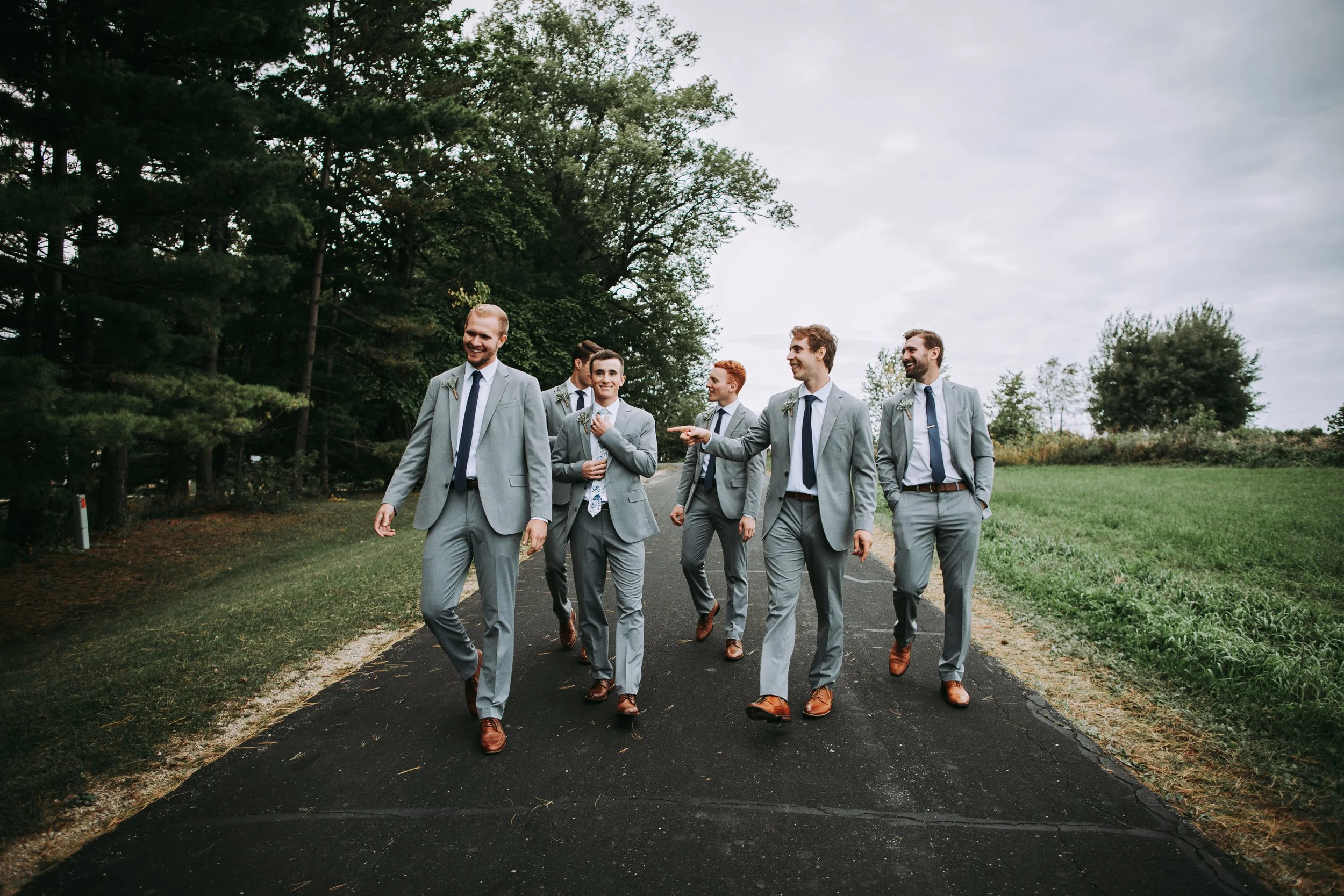 A group of six men in matching light gray suits, white shirts, and dark ties walking outdoors on a paved path, surrounded by trees and grassy areas, with one man smiling and gesturing as others talk and laugh.