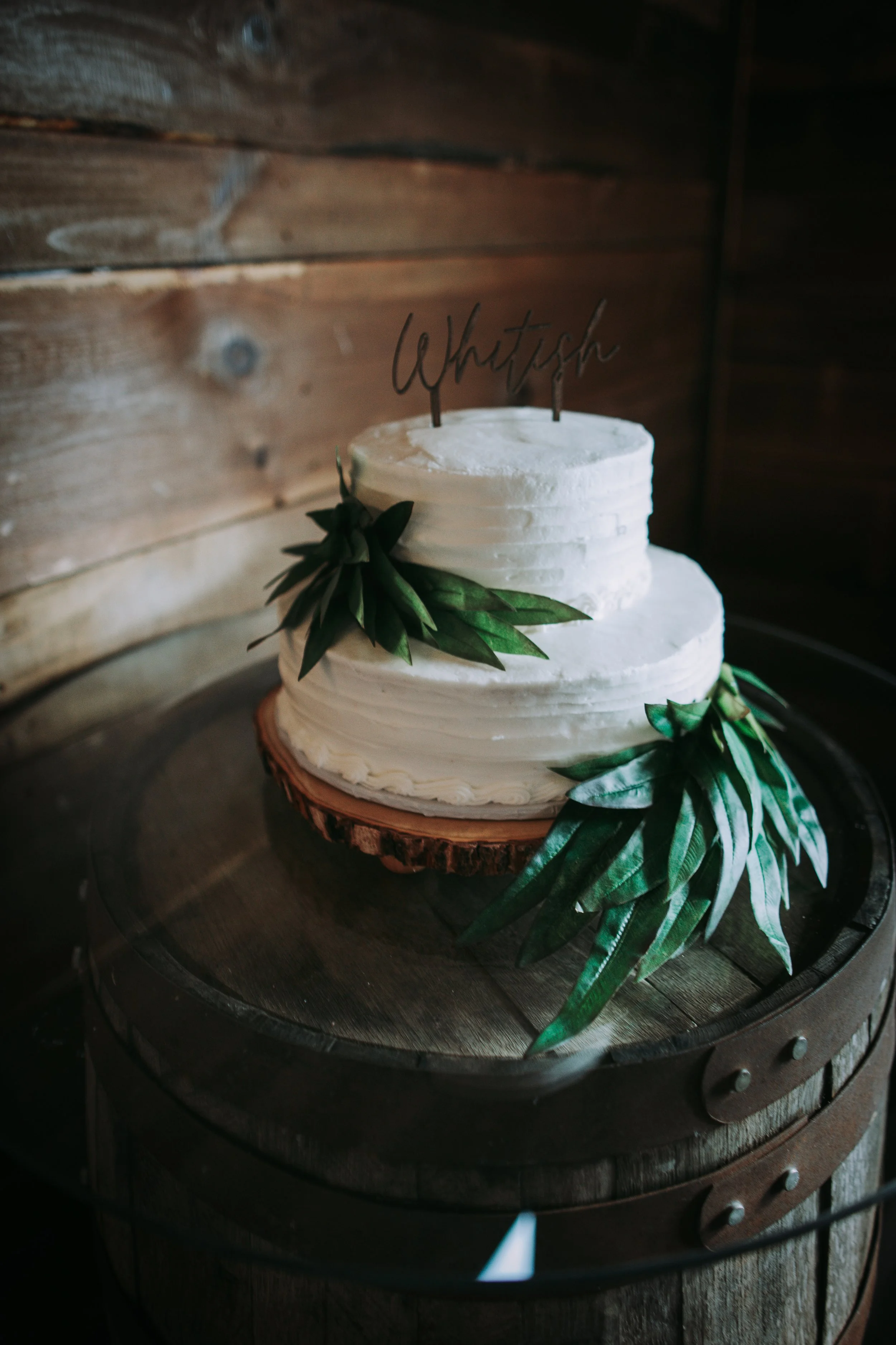 A white wedding cake decorated with green leaves and a topper with the name 'Whitish' in a cursive font, placed on a wooden cake stand.