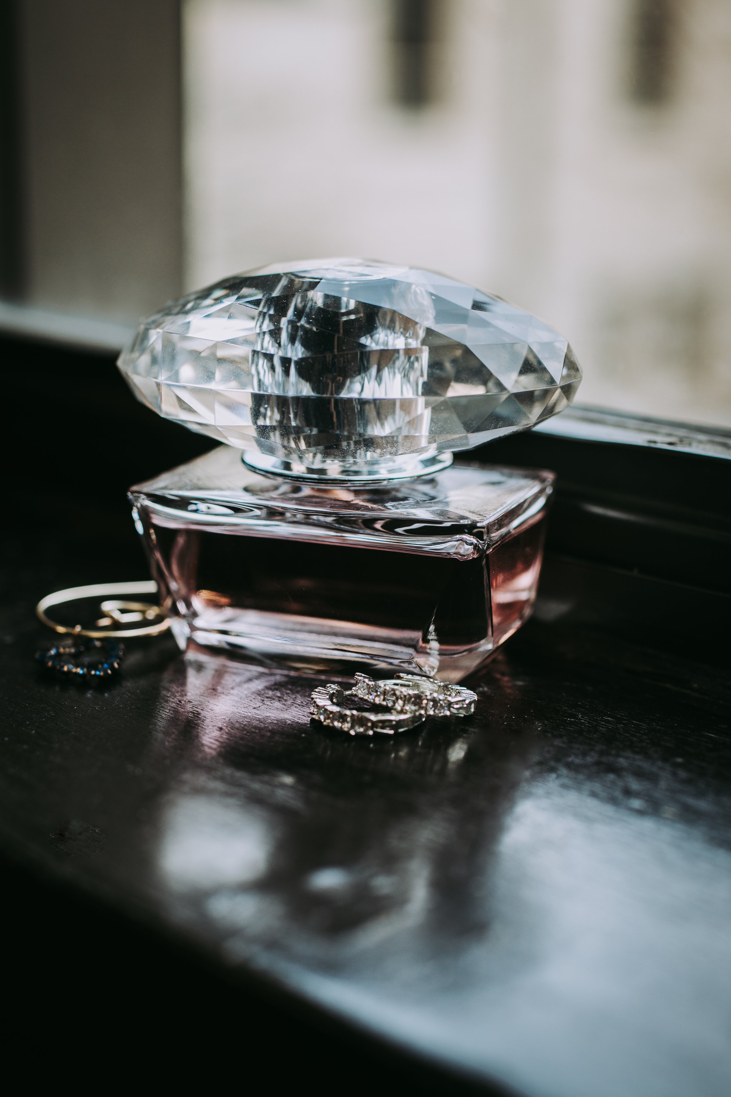 Close-up of a faceted glass perfume bottle with pink liquid, placed on a dark surface alongside jewelry including rings and earrings.