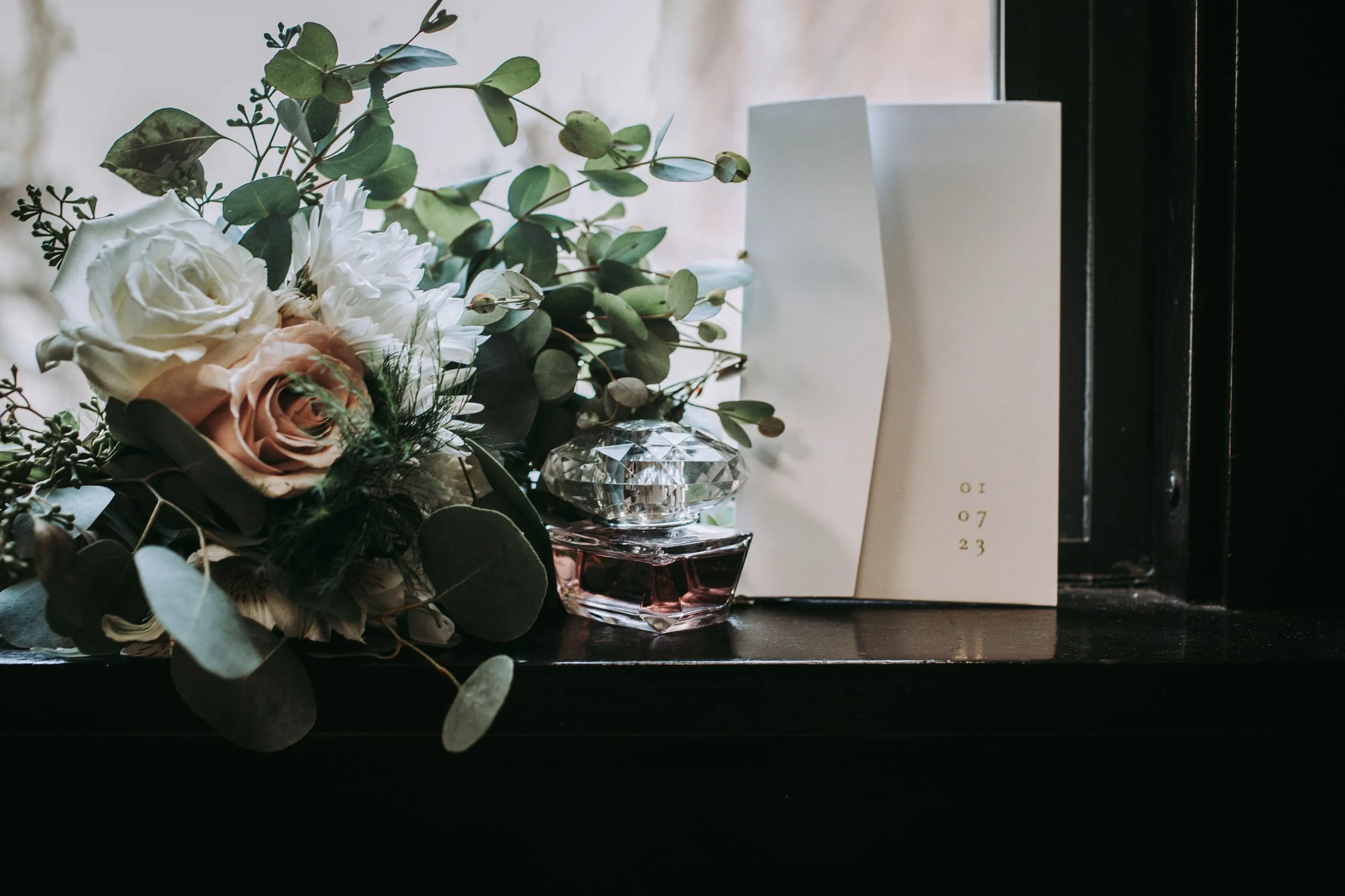 A bouquet of white and peach roses with green eucalyptus leaves, a crystal perfume bottle, and a white card with gold lettering on a black windowsill.