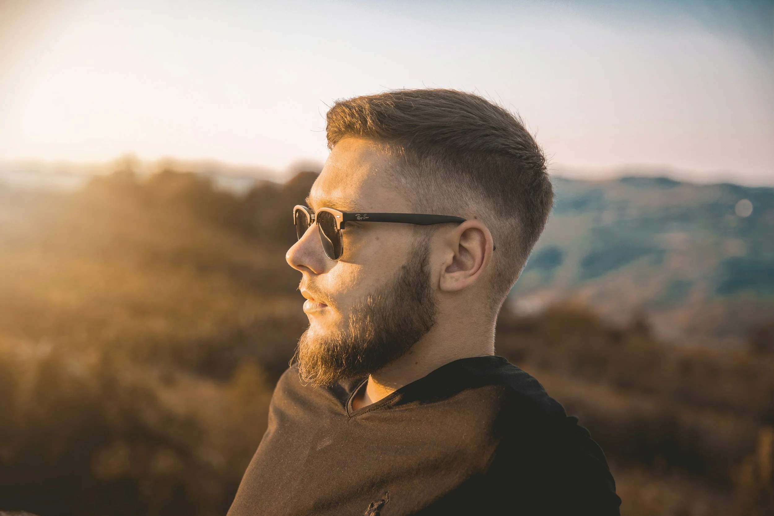 A young man with a beard and short hair wearing sunglasses looking to the side during sunset with a blurred outdoor landscape in the background.
