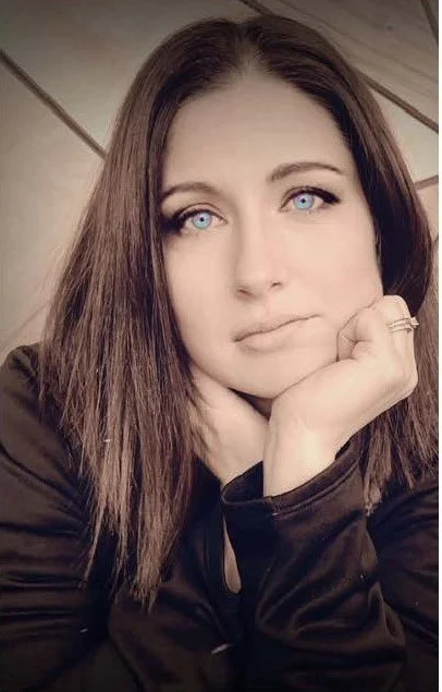 A woman with long brown hair and piercing blue eyes resting her chin on her hand, wearing a black jacket and ring, with a wooden background.