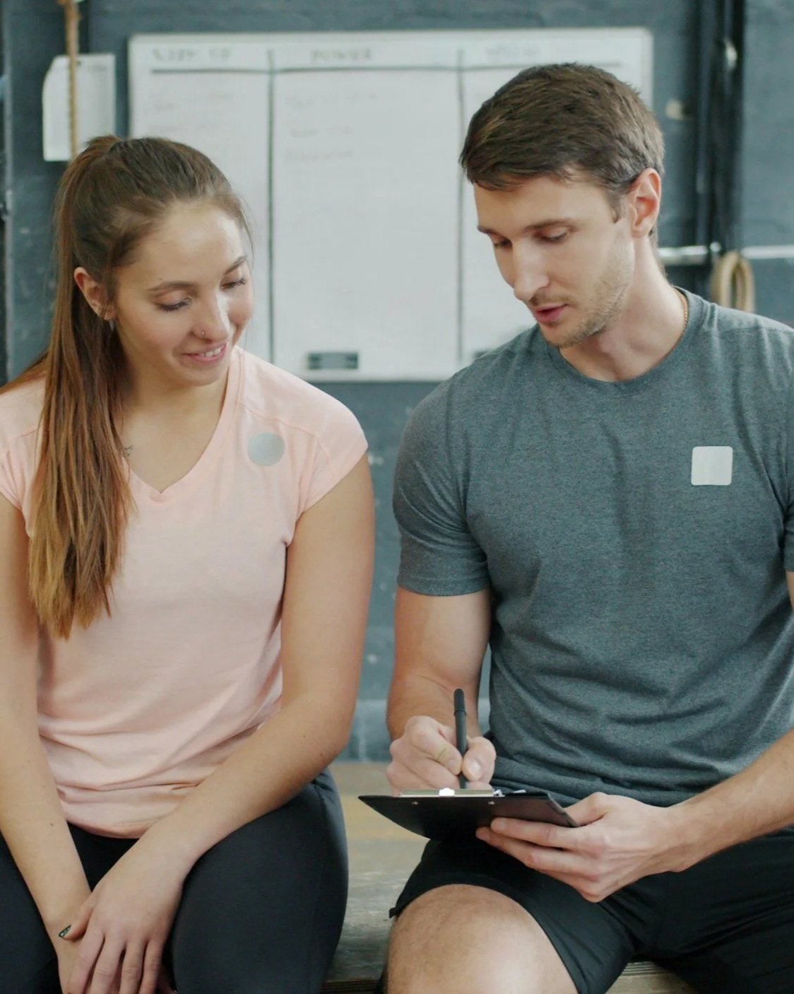 A young woman and a young man sitting closely together at a gym, with the man writing on a clipboard, and the woman smiling at him.