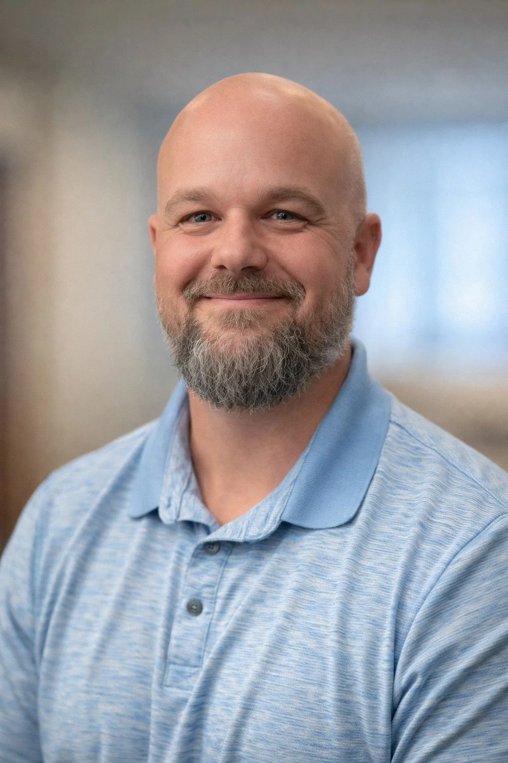 A smiling man with a bald head, grey beard, and blue eyes, wearing a light blue polo shirt, standing in an indoor setting.