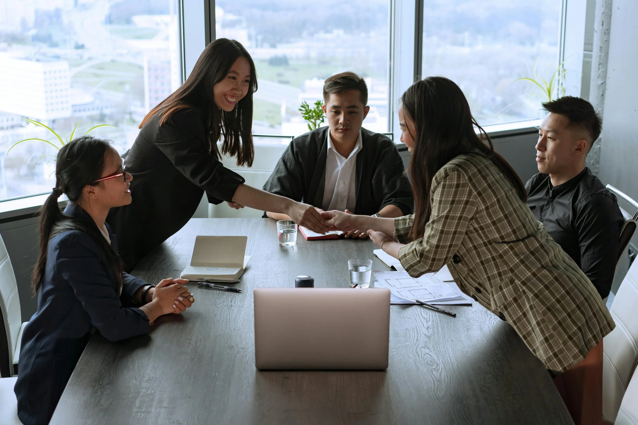 A woman in business attire shaking hands with another woman at a meeting table, while three other people look on in a modern office with large windows and a city view.
