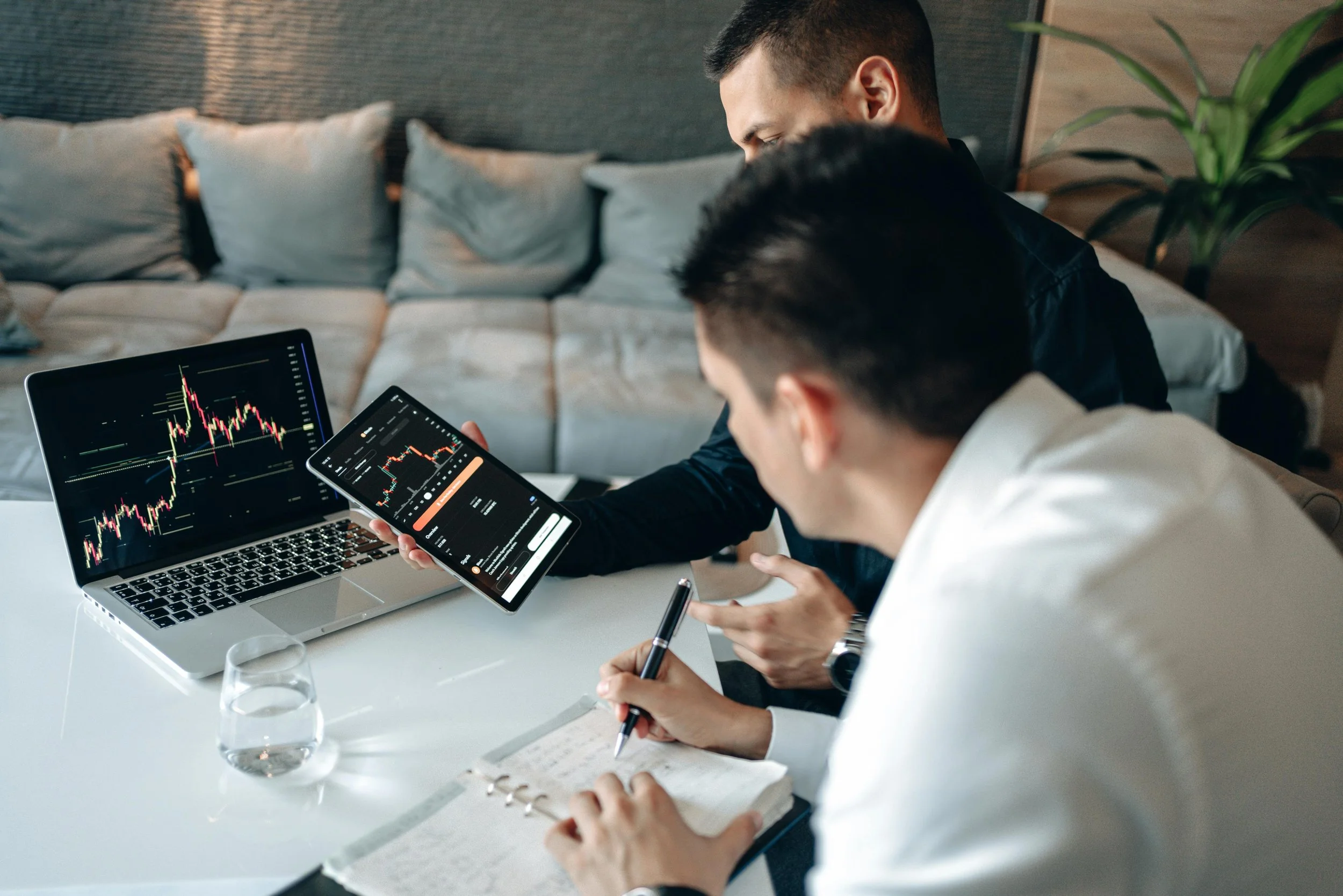 Two men analyzing stock market charts on a laptop and tablet in a modern office setting.