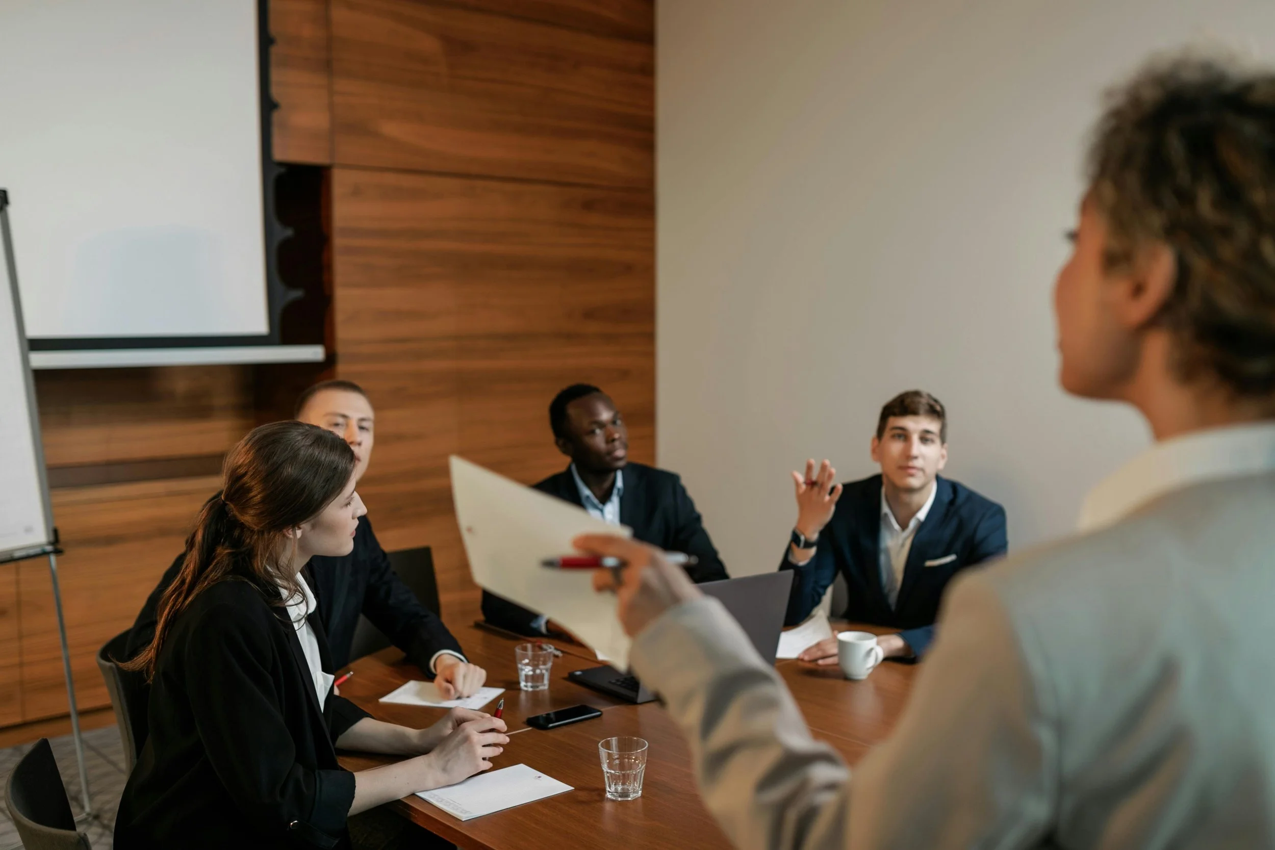 A team of five professionals participating in a meeting around a wooden conference table, with one person standing and speaking while holding papers, and others listening attentively.