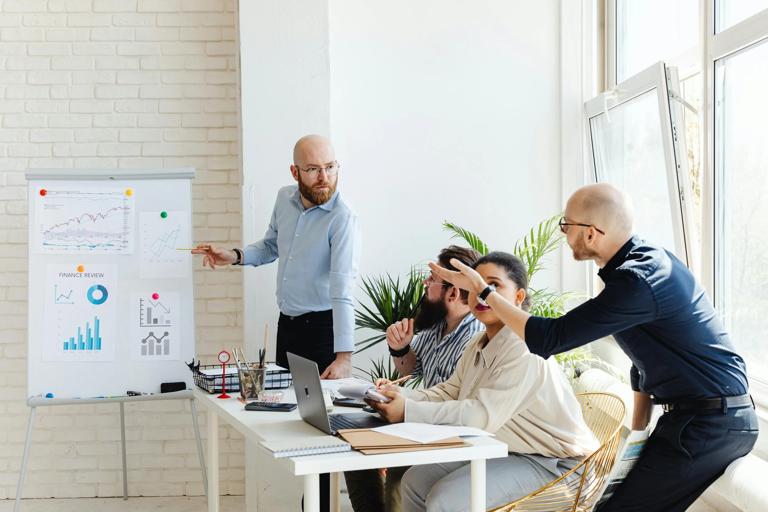 Business team in a meeting room, with a man presenting charts on a whiteboard, while three colleagues listen and discuss, with large windows and plants in the background.