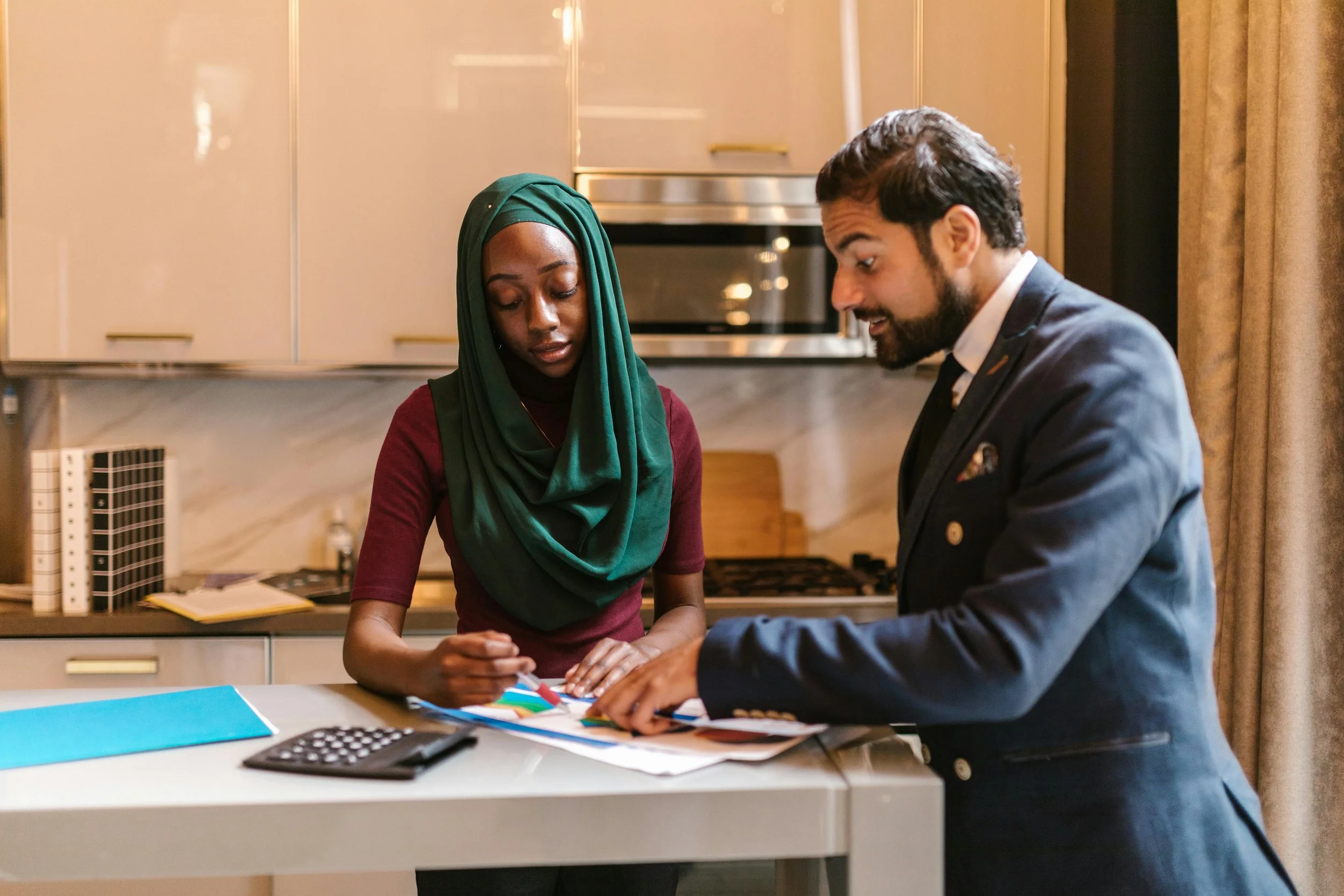 A woman wearing a green hijab and a burgundy top and a man in a navy suit with a beard are standing at a kitchen counter, looking at documents and discussing.