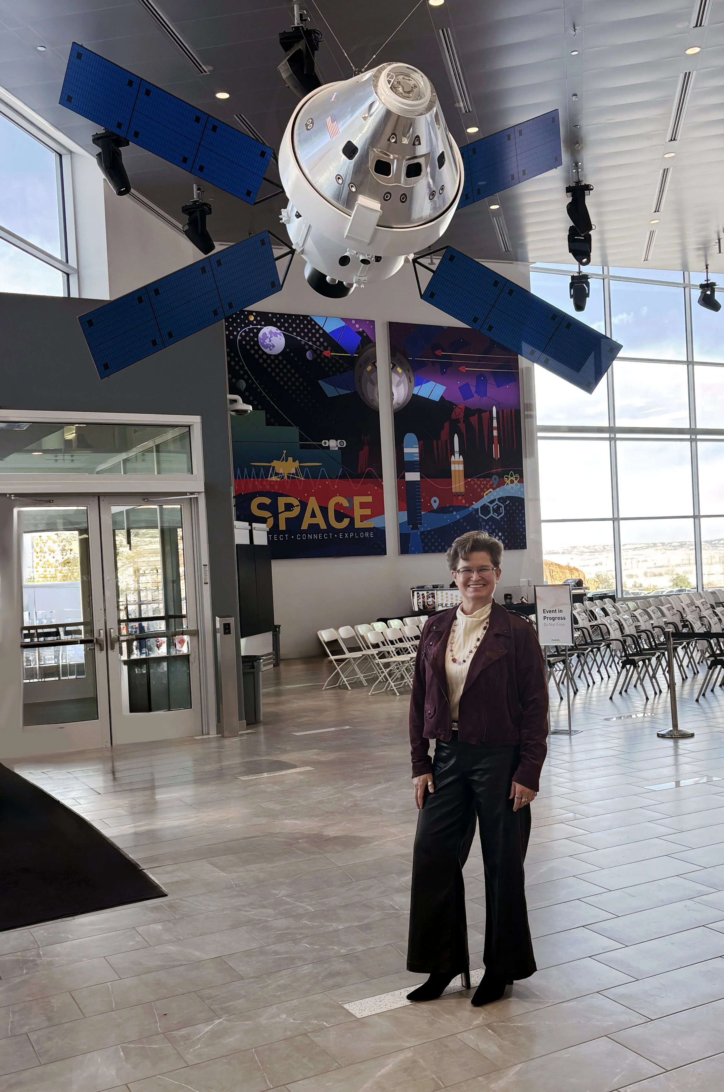 A woman standing indoors in front of a space-themed poster and a large model of a spacecraft with solar panels hanging from the ceiling.