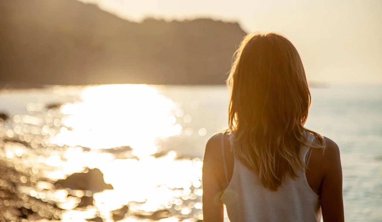 Woman with long hair standing on a rocky beach at sunset, facing the water.