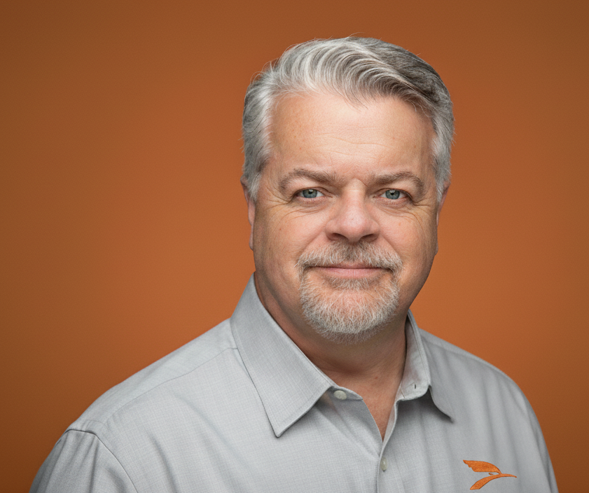 A middle-aged man with light gray hair, a mustache, and beard, wearing a light gray button-up shirt, smiling, against a plain orange background.