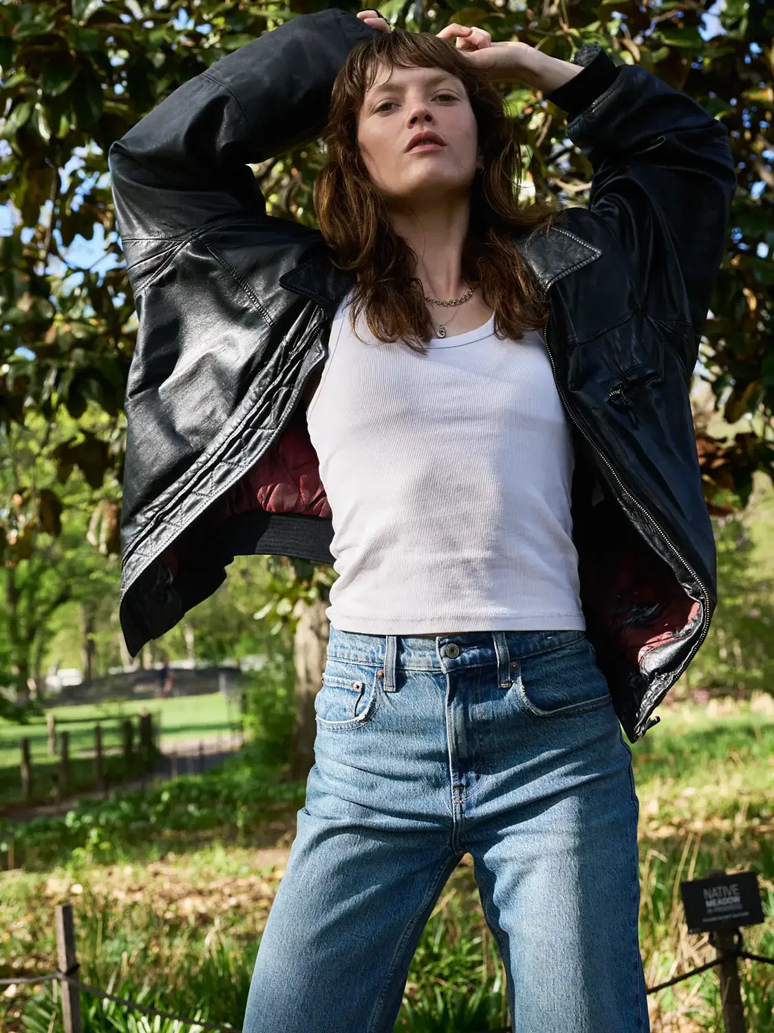 A woman in a white tank top and vintage blue jeans stands outdoors with her arms raised, wearing a black leather jacket. She has shoulder-length brown hair and is looking directly at the camera. Trees and greenery are visible in the background.