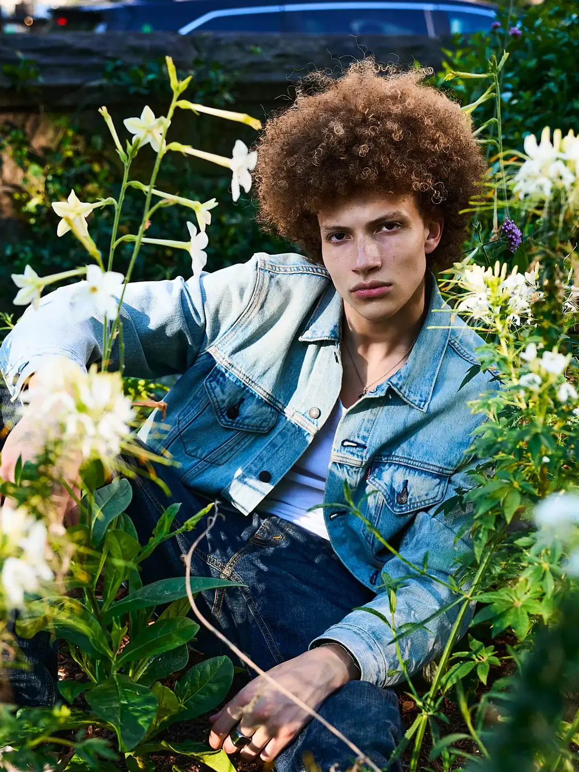 Young person with curly hair wearing denim jacket and jeans surrounded by flowering plants outdoors.