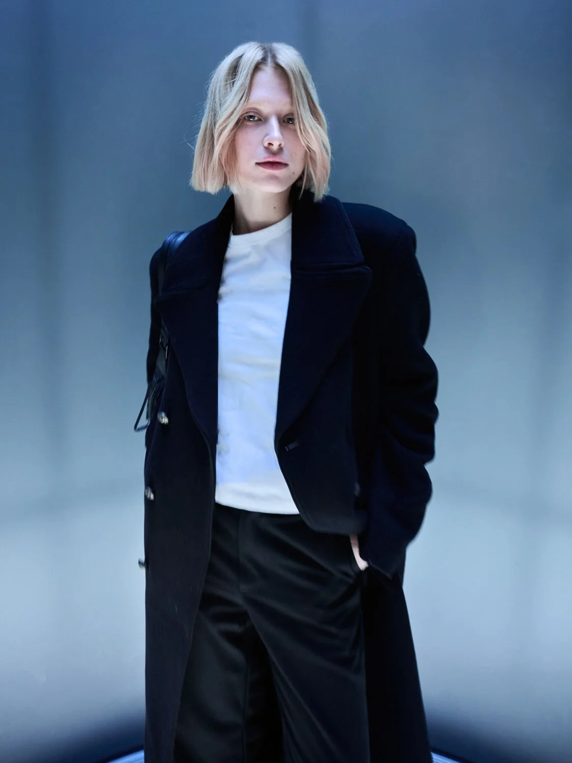 A young woman with shoulder-length blond hair stands against a plain gray background, wearing a black coat, white shirt, black pants, and carrying a black backpack.