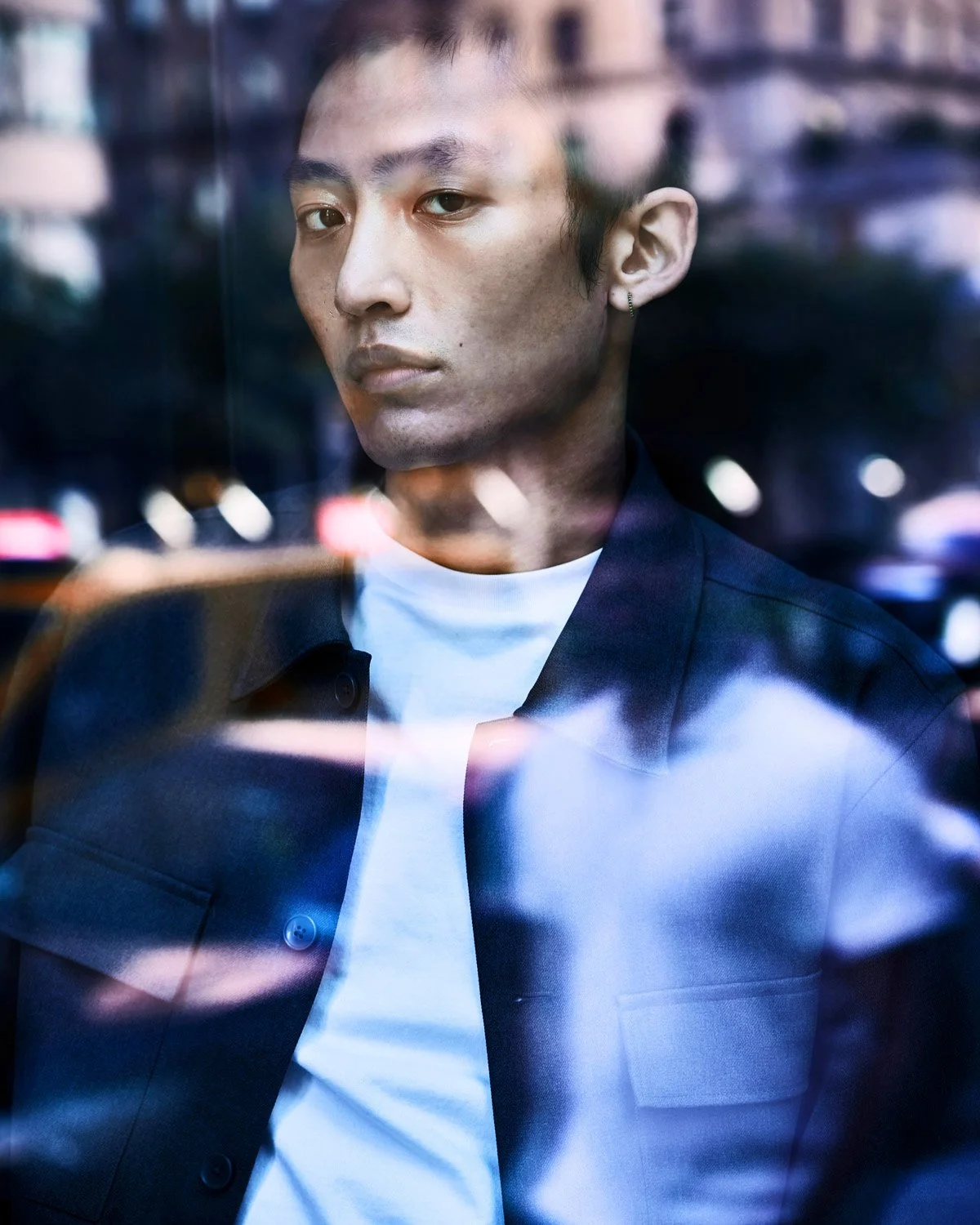 A young man with short hair and an earring looking through a glass window in a city at night, with reflections of lights on the glass.