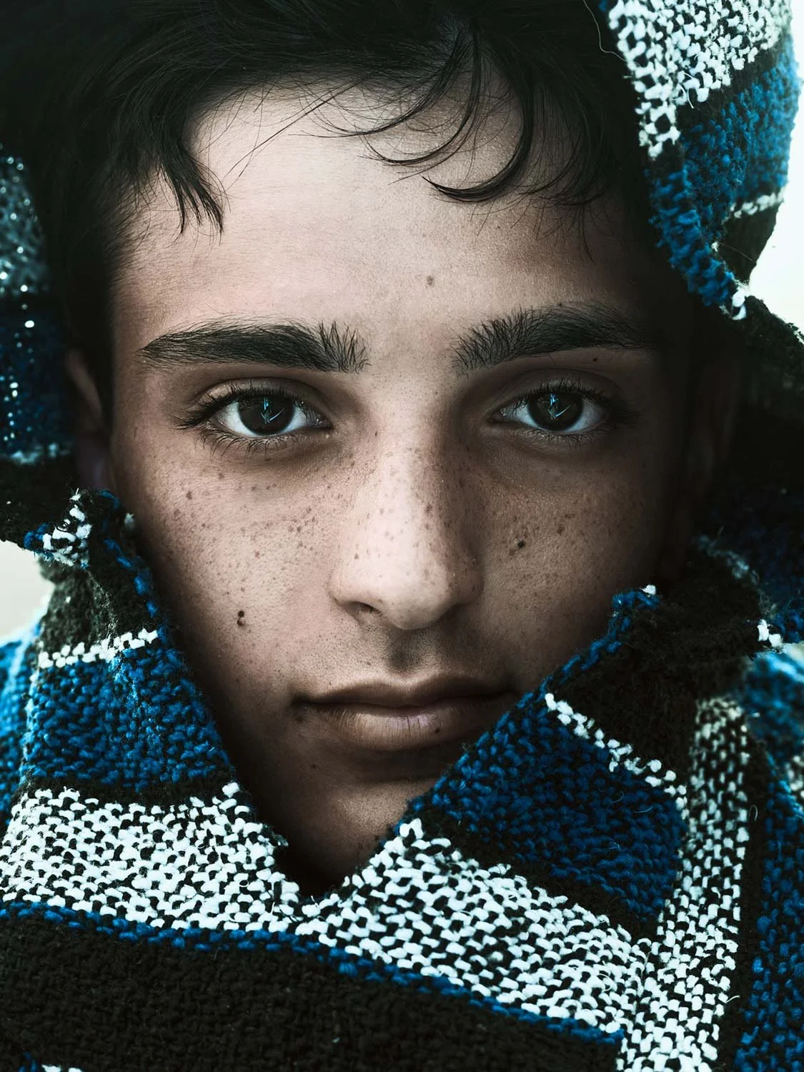 Close-up of a young person with blue eyes and dark hair, wrapped in a blue and black plaid blanket.
