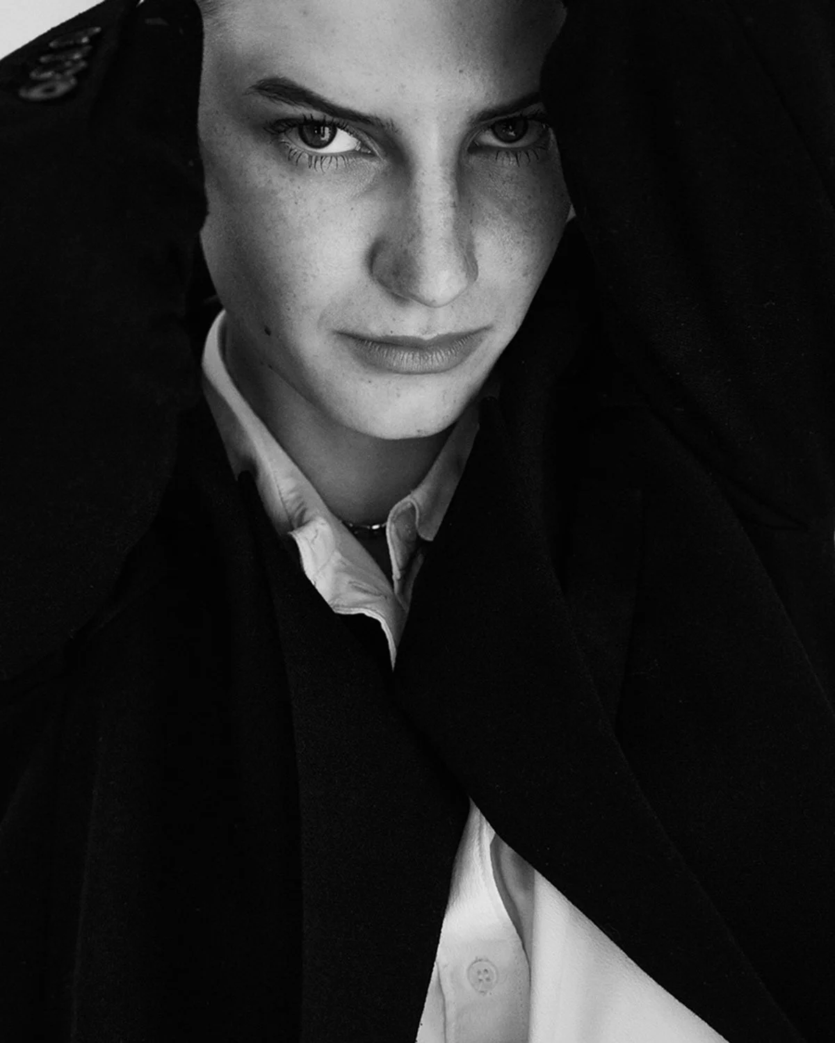 A black and white close-up portrait of a woman looking intensely at the camera with her hands on her head, wearing a white shirt and a dark blazer.