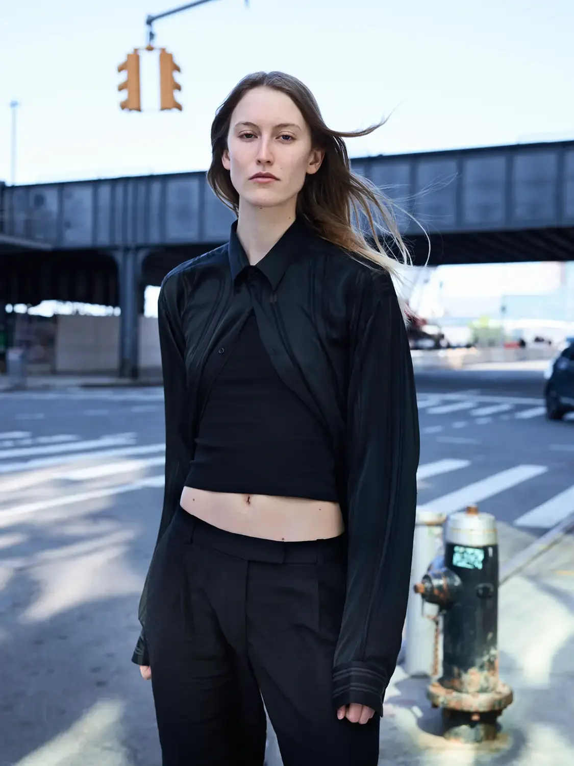 Young woman with long hair standing on city street near a fire hydrant, wearing a black crop top, pants, and a black jacket against a background of overpass and traffic.