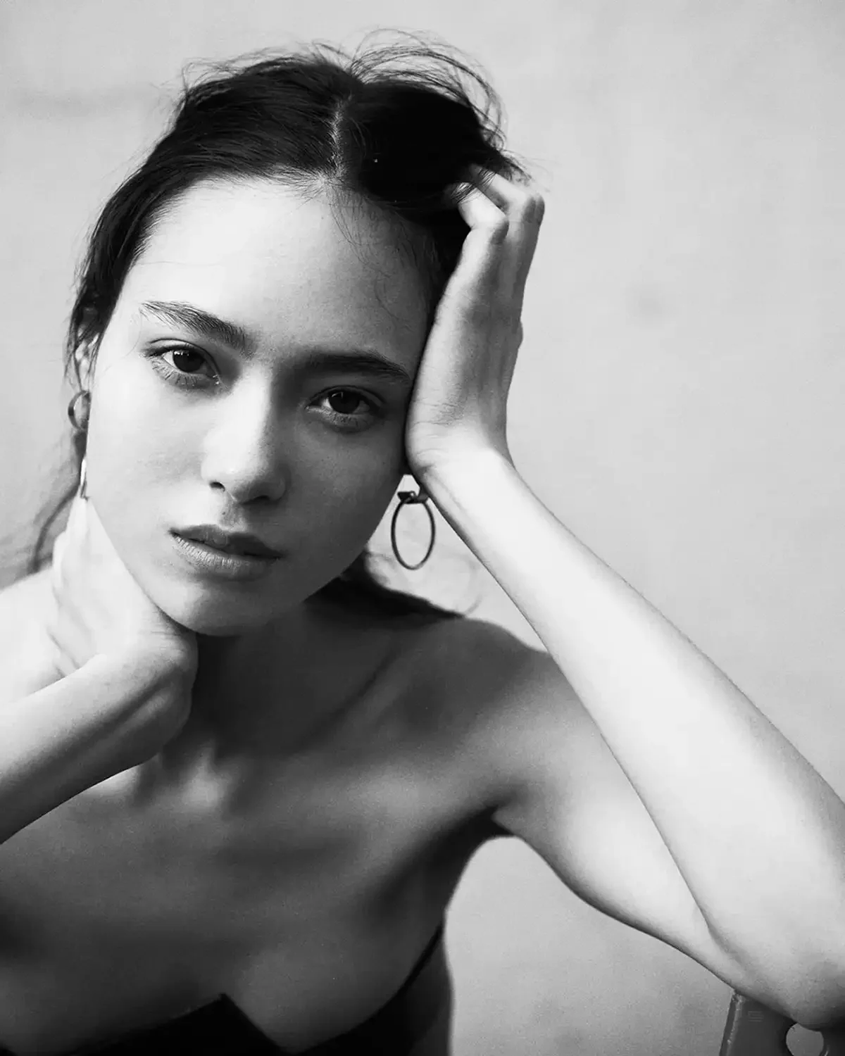 Black and white portrait of a young woman with dark hair, one hand resting on her head, wearing a single hoop earring and a strapless top.