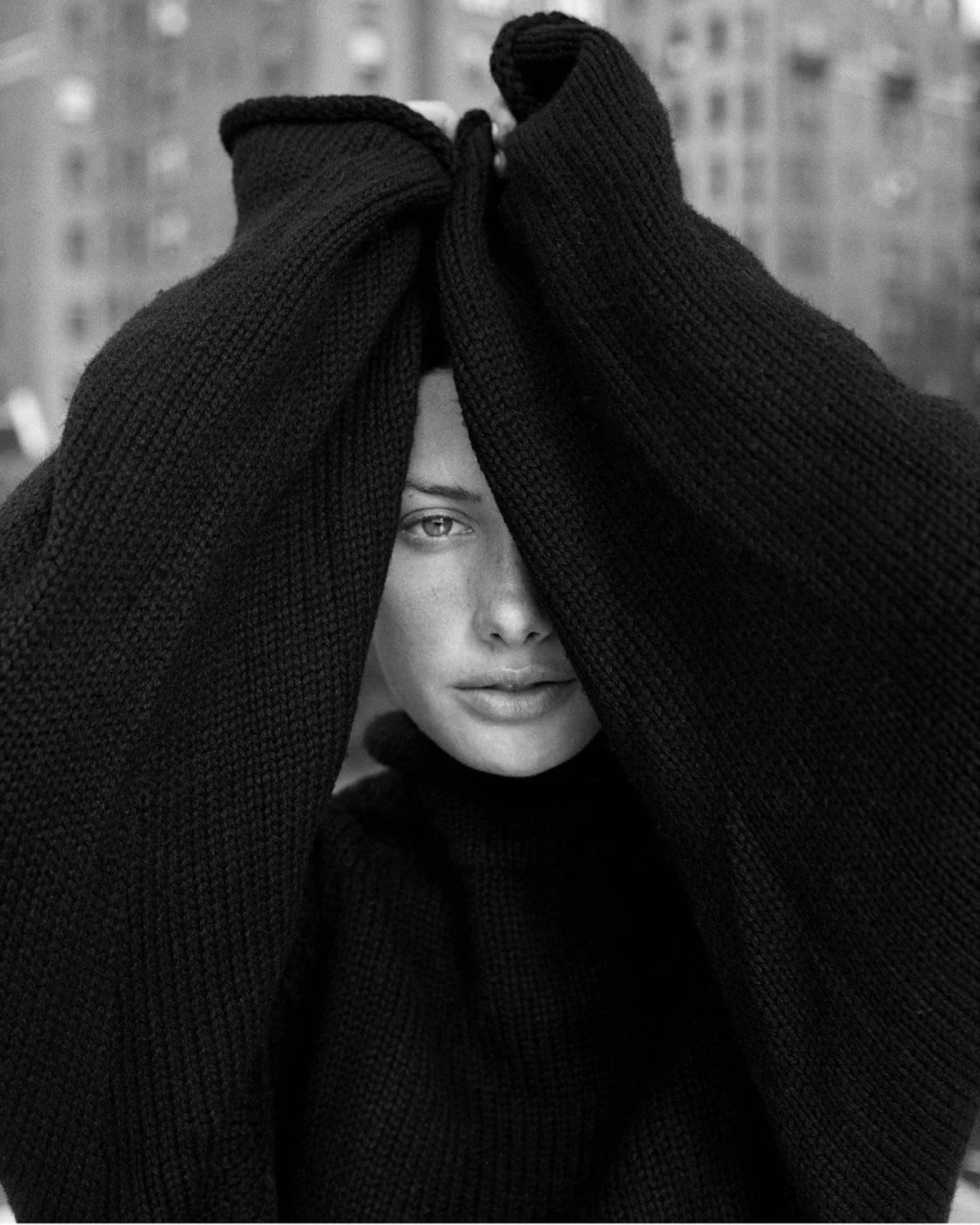 Close-up black and white portrait of a woman with half her face partially covered by overlapping black knitted sleeves, outdoors with a blurred urban background.