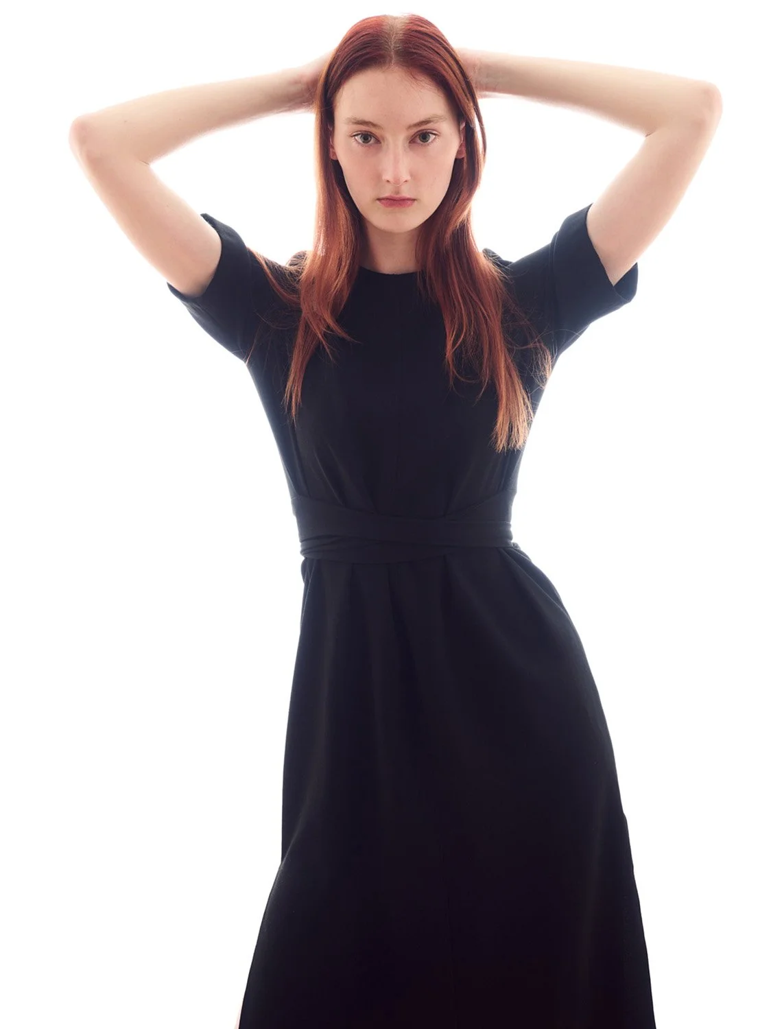 A woman with long red hair wearing a black dress, standing with her arms raised behind her head against a white background.