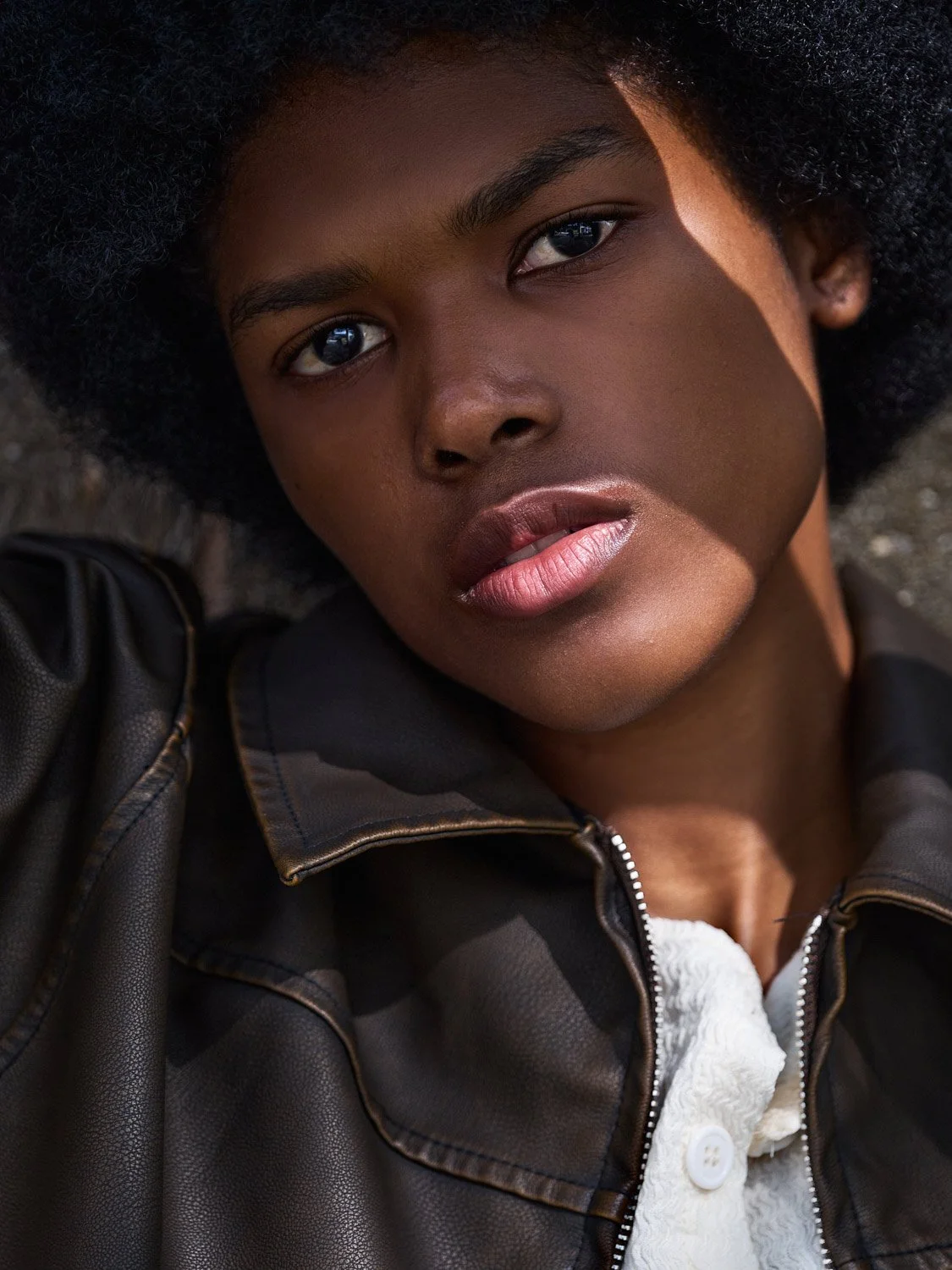 Beauty shot, close-up of a woman with dark skin and curly hair, lying on the ground, wearing a dark brown leather jacket and a white textured shirt.