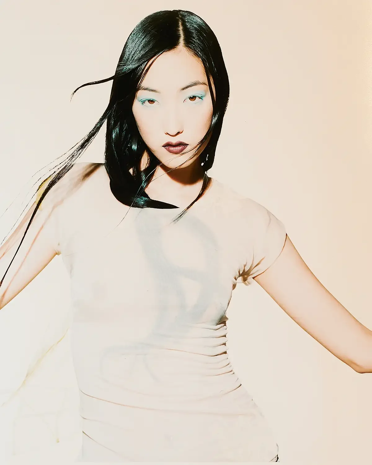 A woman with long black hair, wearing makeup, a light-colored top with short sleeves, stands against a plain background, striking a confident pose.