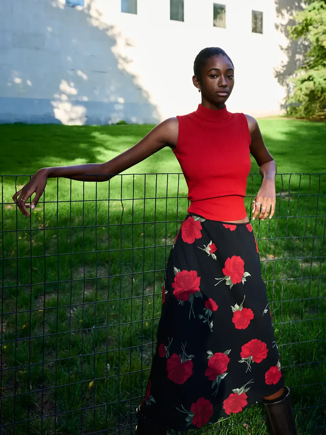 A woman wearing a red sleeveless turtleneck top and a black skirt with large red roses, standing outdoors, leaning on a metal fence with one arm, with a grassy area and modern building in the background.