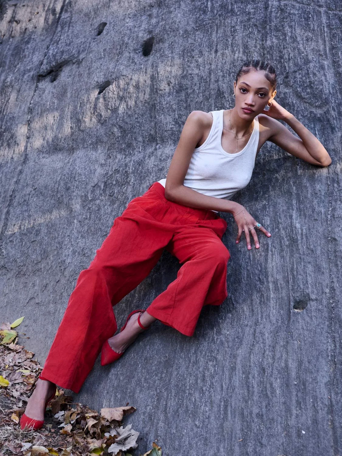 A young woman with braided hair, wearing a white tank top, red pants, and red high heels, reclining on a large rock or stone surface outdoors, surrounded by fallen leaves.