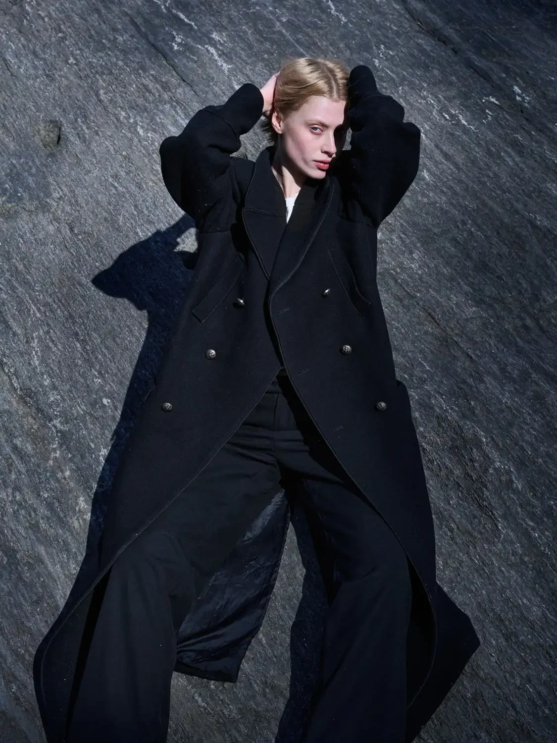 A woman with blonde hair lies on a dark, textured surface, wearing a black coat and black pants, with her hands behind her head and looking at the camera.