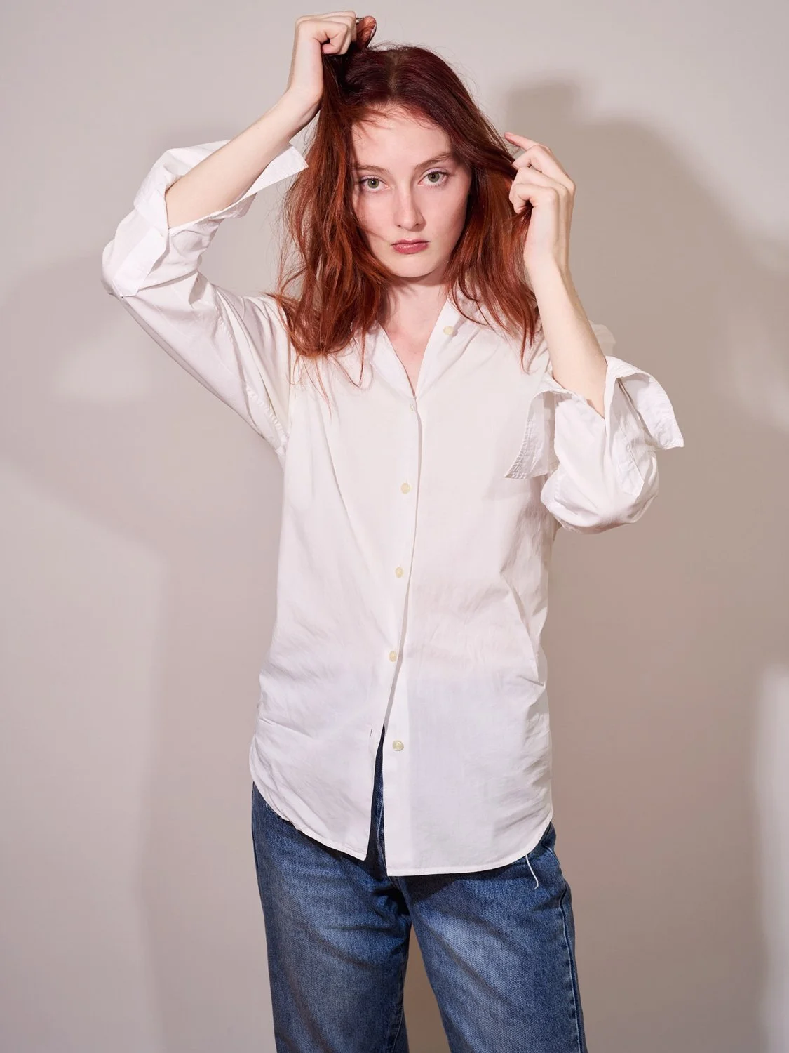 A young woman with long red hair wearing a white button-up shirt and blue jeans, standing against a plain background, touching her hair and looking into the camera.