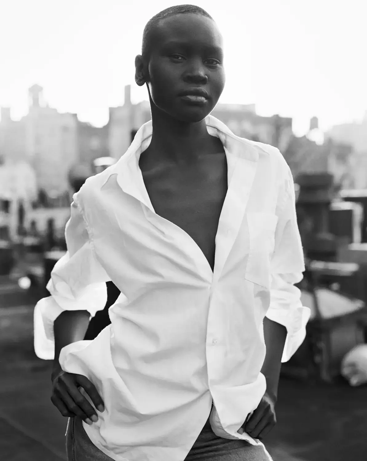 A black and white photo of Alek Wek standing outdoors on a rooftop with city buildings in the background. She is wearing a white button-down shirt with the top few buttons undone.