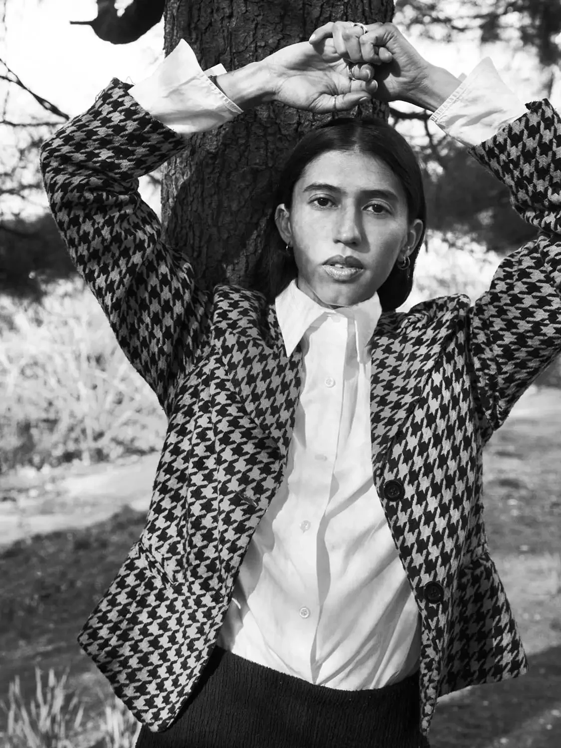 A woman with medium skin tone and dark hair standing outdoors, leaning against a tree with her arms raised above her head, gripping onto the trunk. She is wearing a patterned blazer, a white shirt, and a dark skirt, and is looking directly at the cam