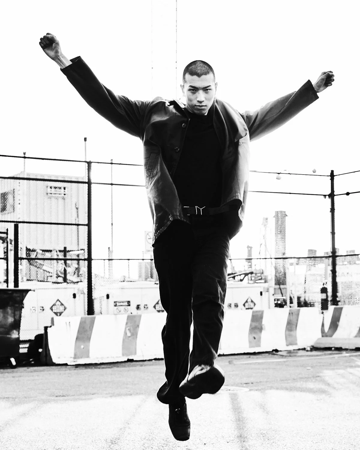 Black and white photo of a man jumping with arms outstretched on an urban rooftop, with city buildings and a chain-link fence in the background.