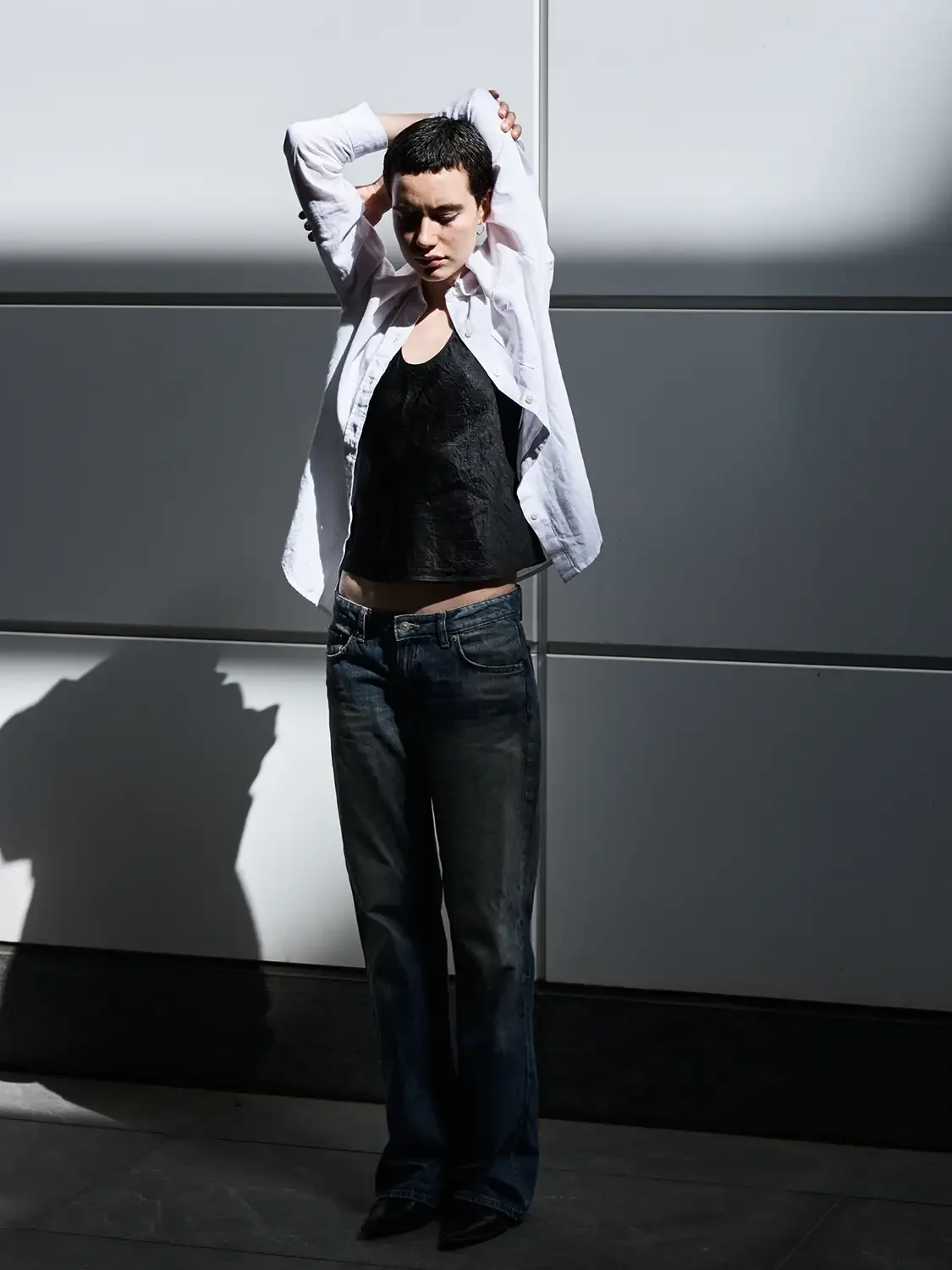 A woman with short dark hair standing against a gray wall with shadows, wearing a white open shirt over a black top and dark jeans, with her arms raised behind her head and eyes closed.