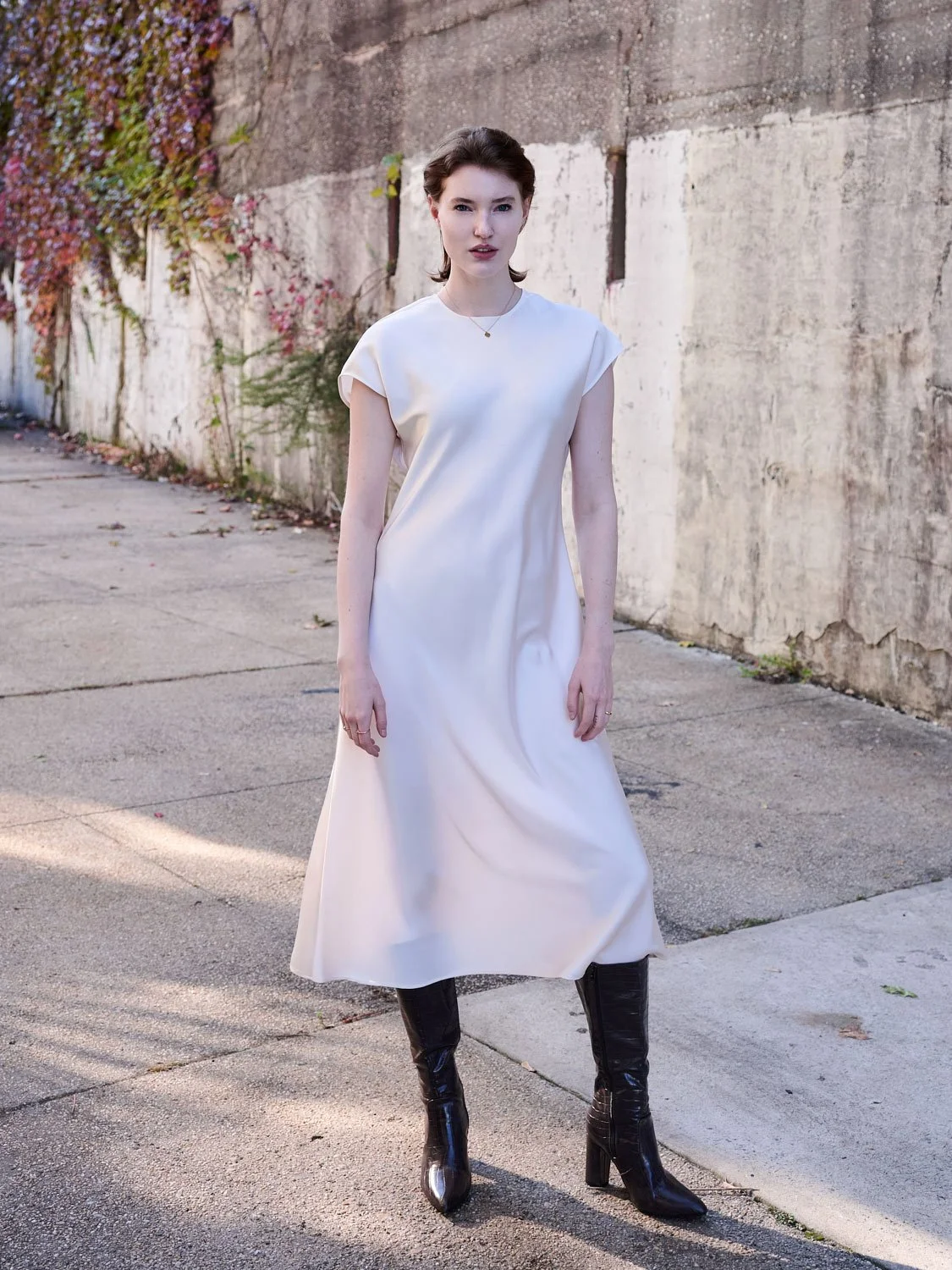 A woman with short brown hair wearing a long white dress and black high-heeled boots standing outdoors on a sidewalk, with a weathered wall and some foliage in the background.
