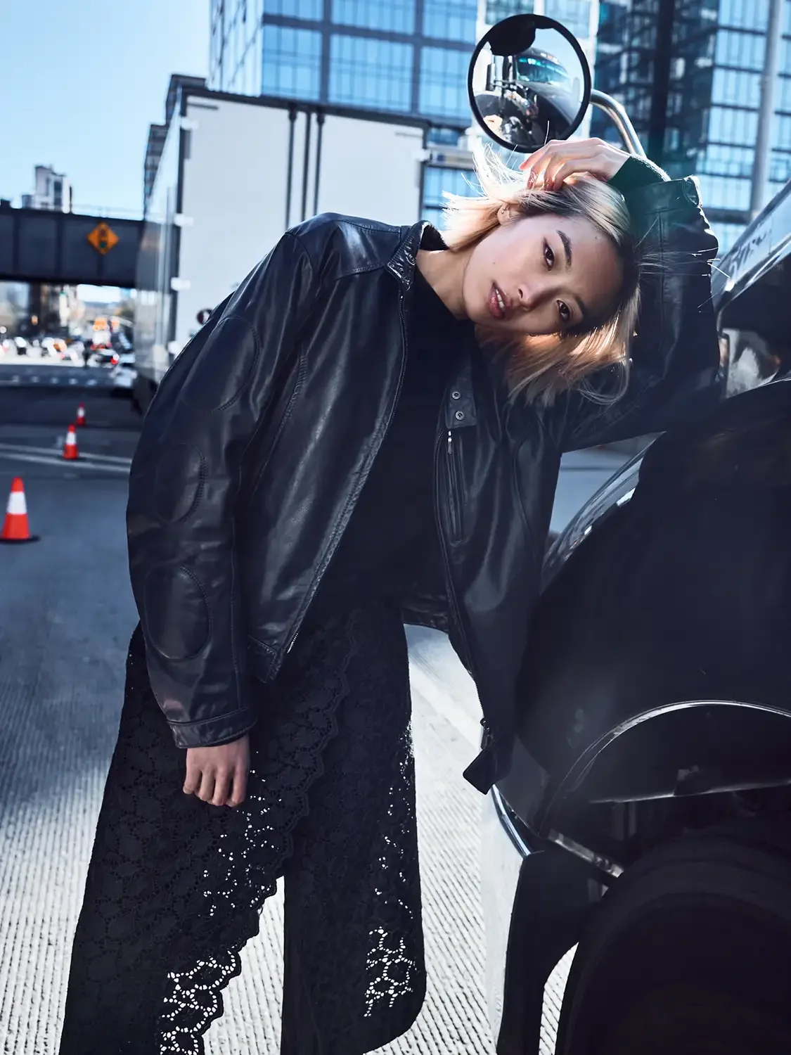 An Asian woman in a black leather jacket and lace pants leaning on a black truck in an urban area with glass buildings and traffic cones.
