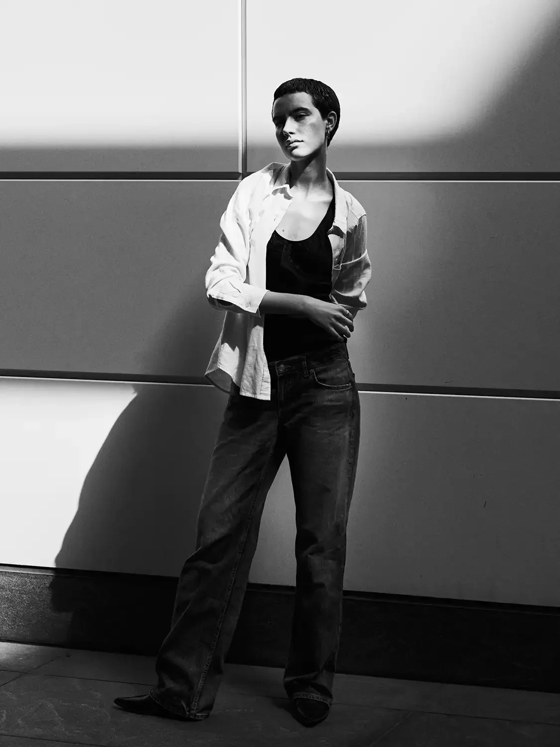 A woman with short hair standing against a wall, wearing a white shirt, black top, and jeans, with sunlight casting shadows on her.