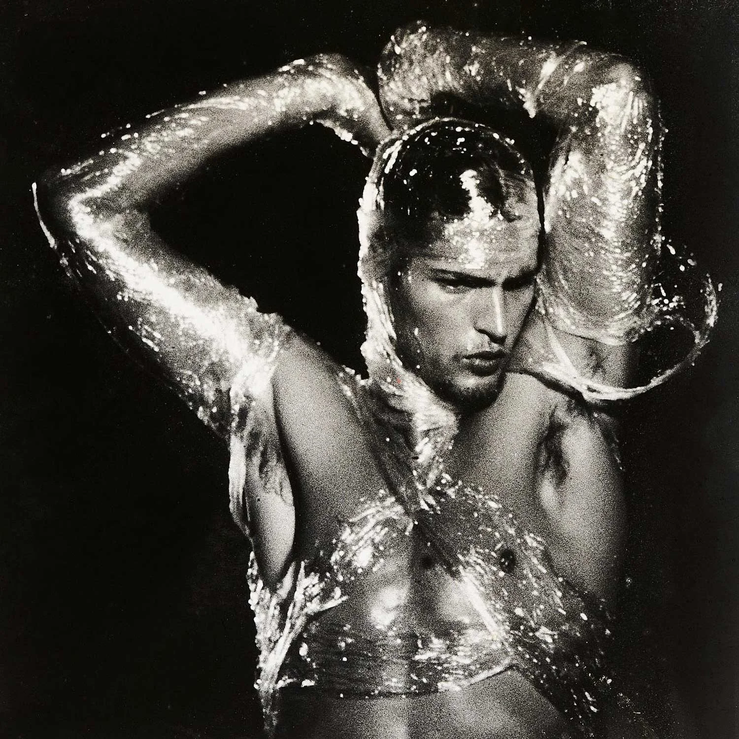 Black and white photograph of a man with slicked-back hair, wearing saran wrap, translucent, puffy sleeves, posing with her arms behind his head.