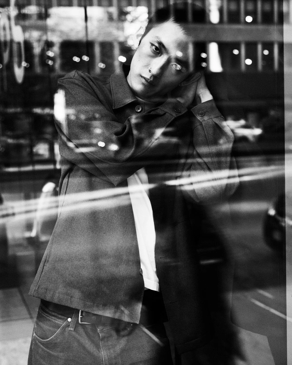 A person leaning against a window, looking contemplative, with reflections and bokeh lights visible in the glass, in black and white.
