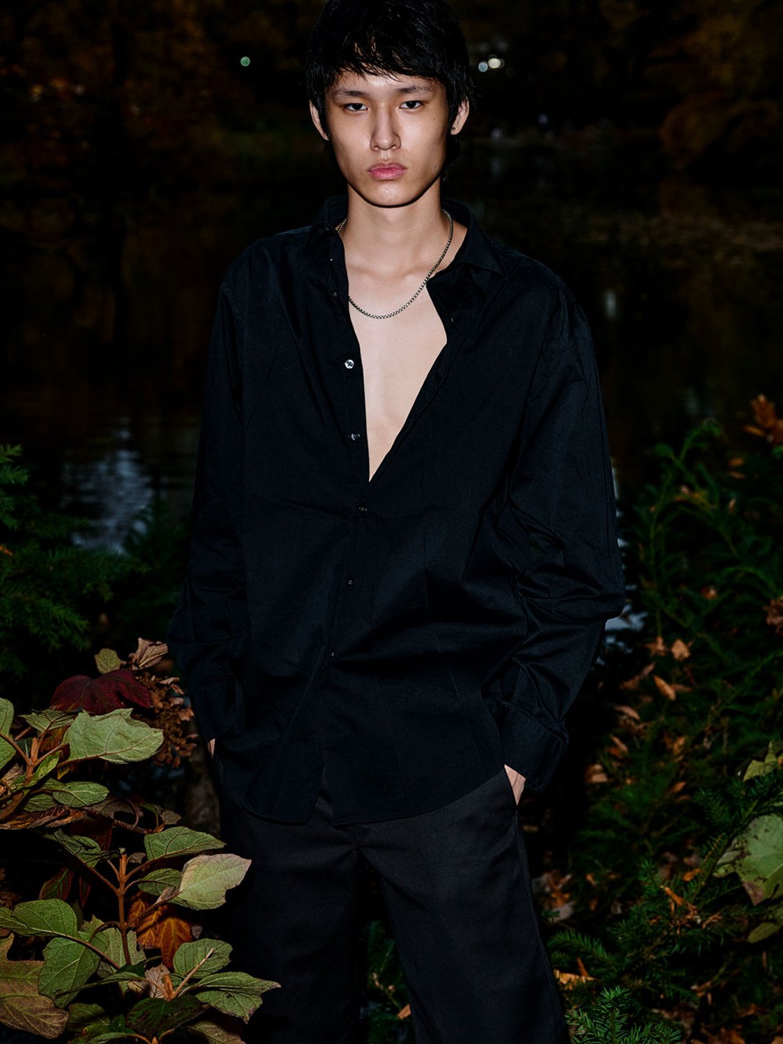 A young man in a black button-up shirt and black pants standing outdoors at night, with hands in pockets, surrounded by green plants and autumn leaves.