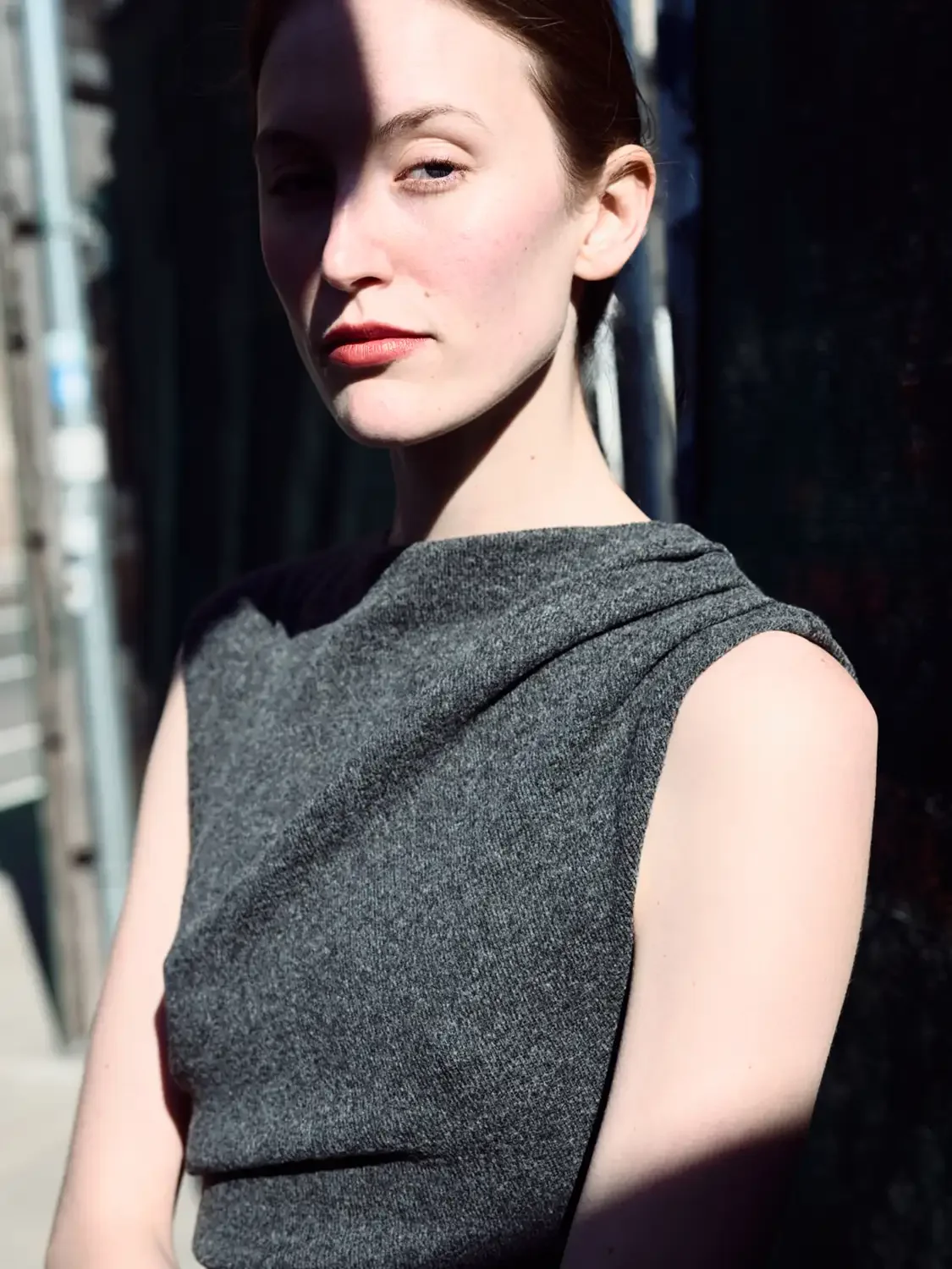 Portrait of a beautiful woman with fair skin and dark hair, wearing a sleeveless gray dress, standing outdoors in bright sunlight.