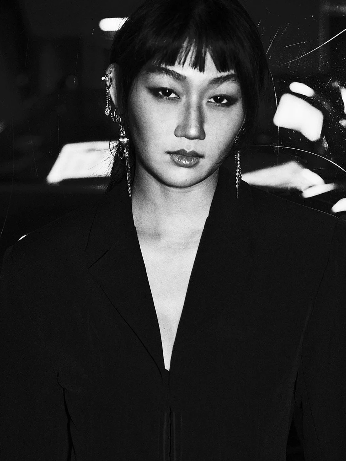 Black and white photo of a woman with short dark hair and earrings, wearing a dark blazer, looking directly at the camera.