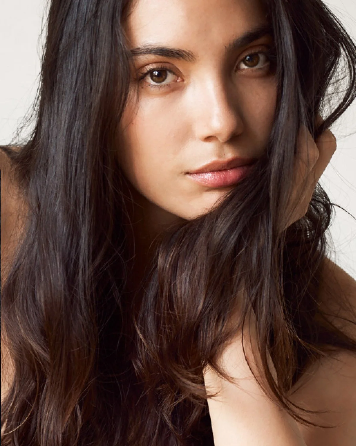 Close-up portrait of a woman with long dark wavy hair, brown eyes, and soft pink lips, looking directly at the camera.