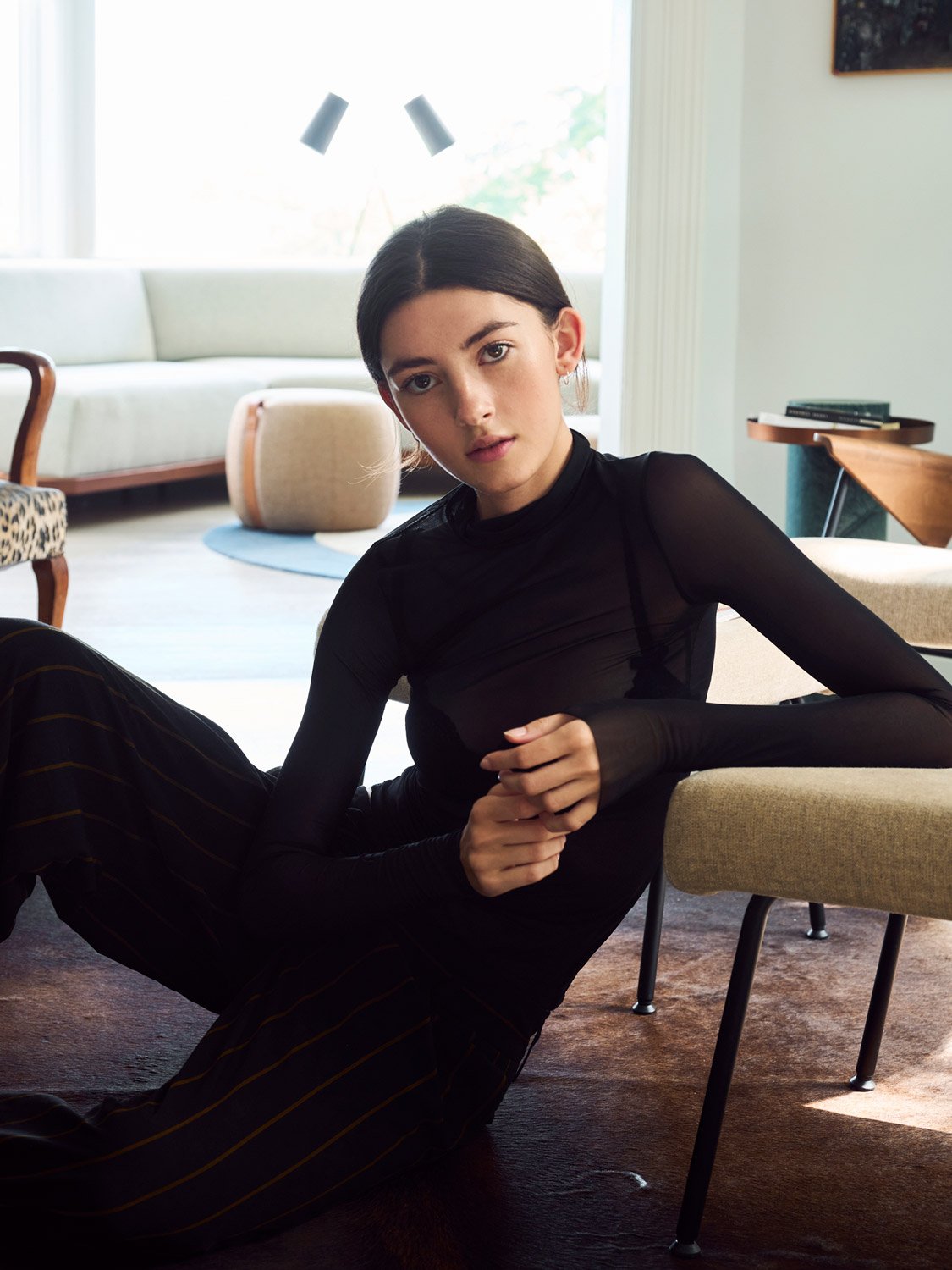 A young woman with dark hair, wearing a black long-sleeve top and striped pants, sitting on the floor in a living room with modern furniture and large windows.