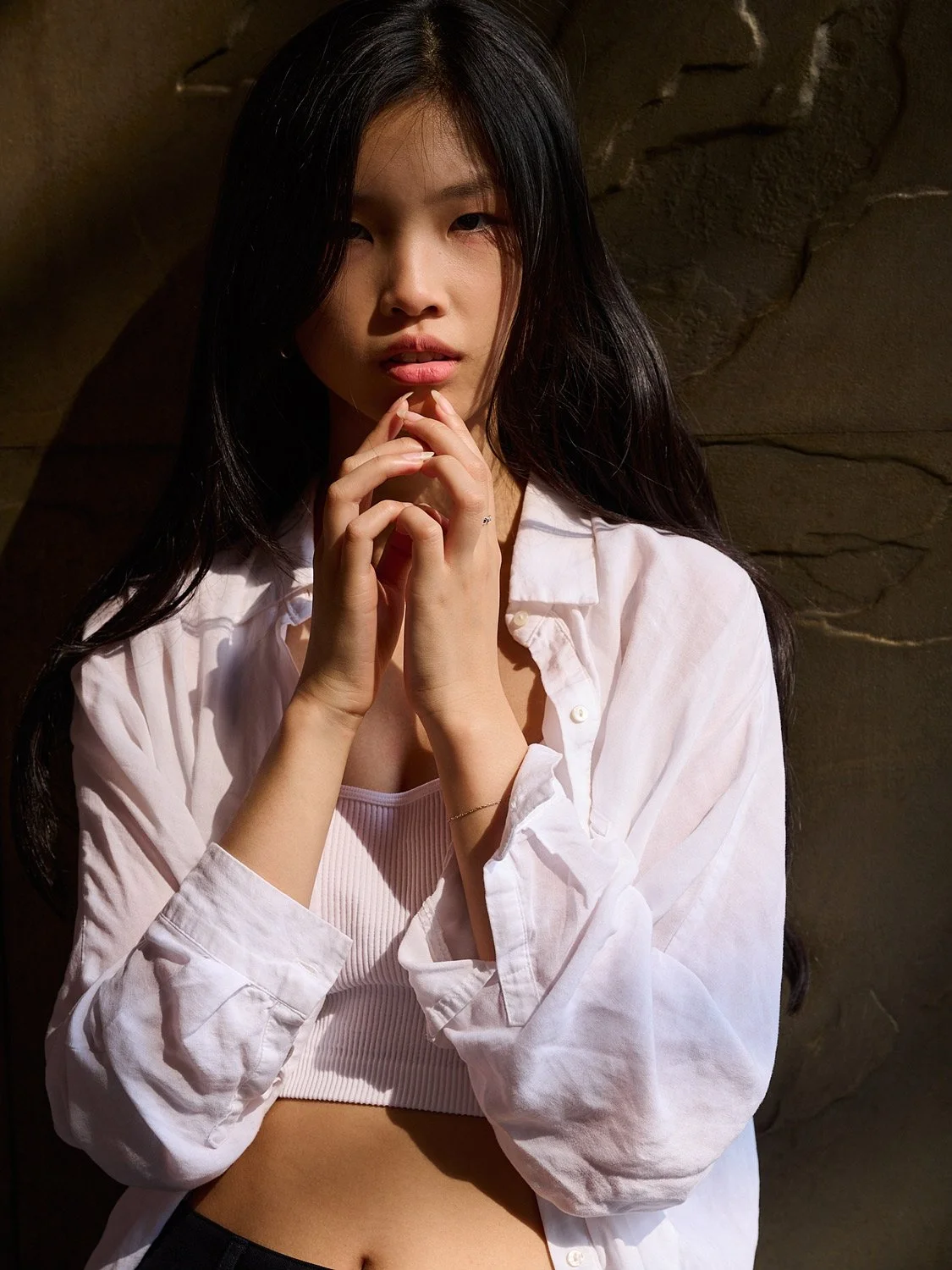 A young woman with long black hair, wearing a white shirt over a pink ribbed tank top, standing against a textured brown stone wall, looking into the camera with her hands clasped together near her chin.